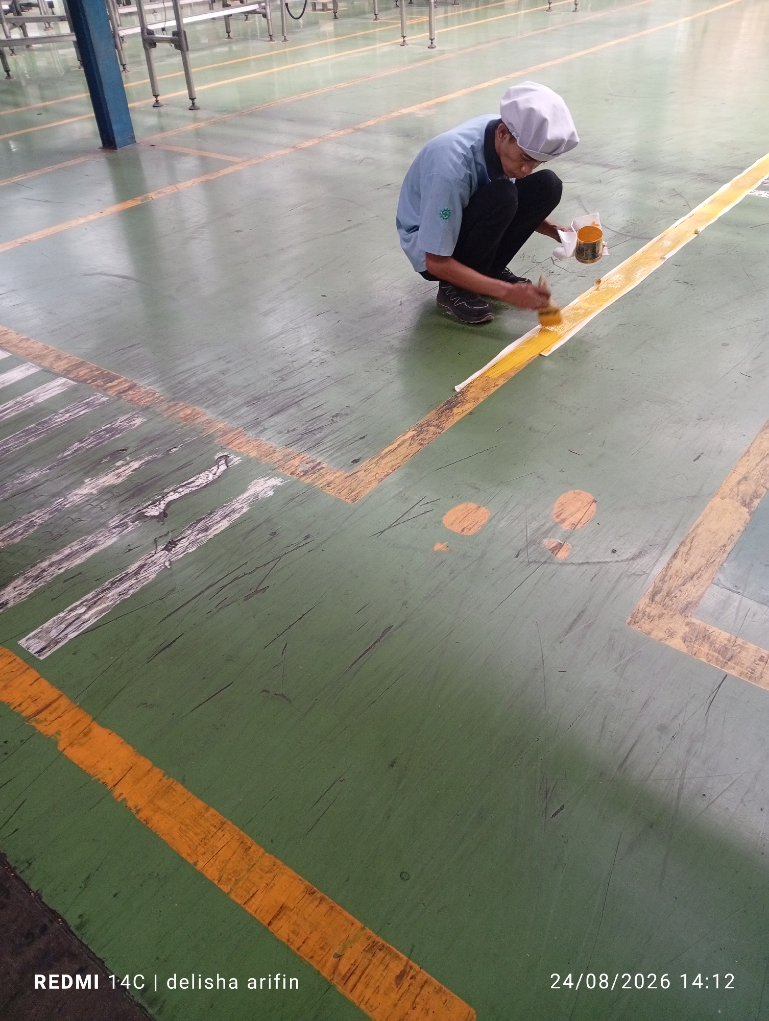 In the city of Gambir Kuning, Indonesia, a man wearing a blue shirt, black pants, and a white cap is kneeling on a green industrial floor. He is holding a small can of yellow paint and a brush, and he is actively painting an orange line on the floor. The floor is marked with other orange and white lines, indicating a designated pathway or work area. The scene appears to be inside a factory or warehouse, with metal structures visible in the background. The lighting suggests it is daytime.
