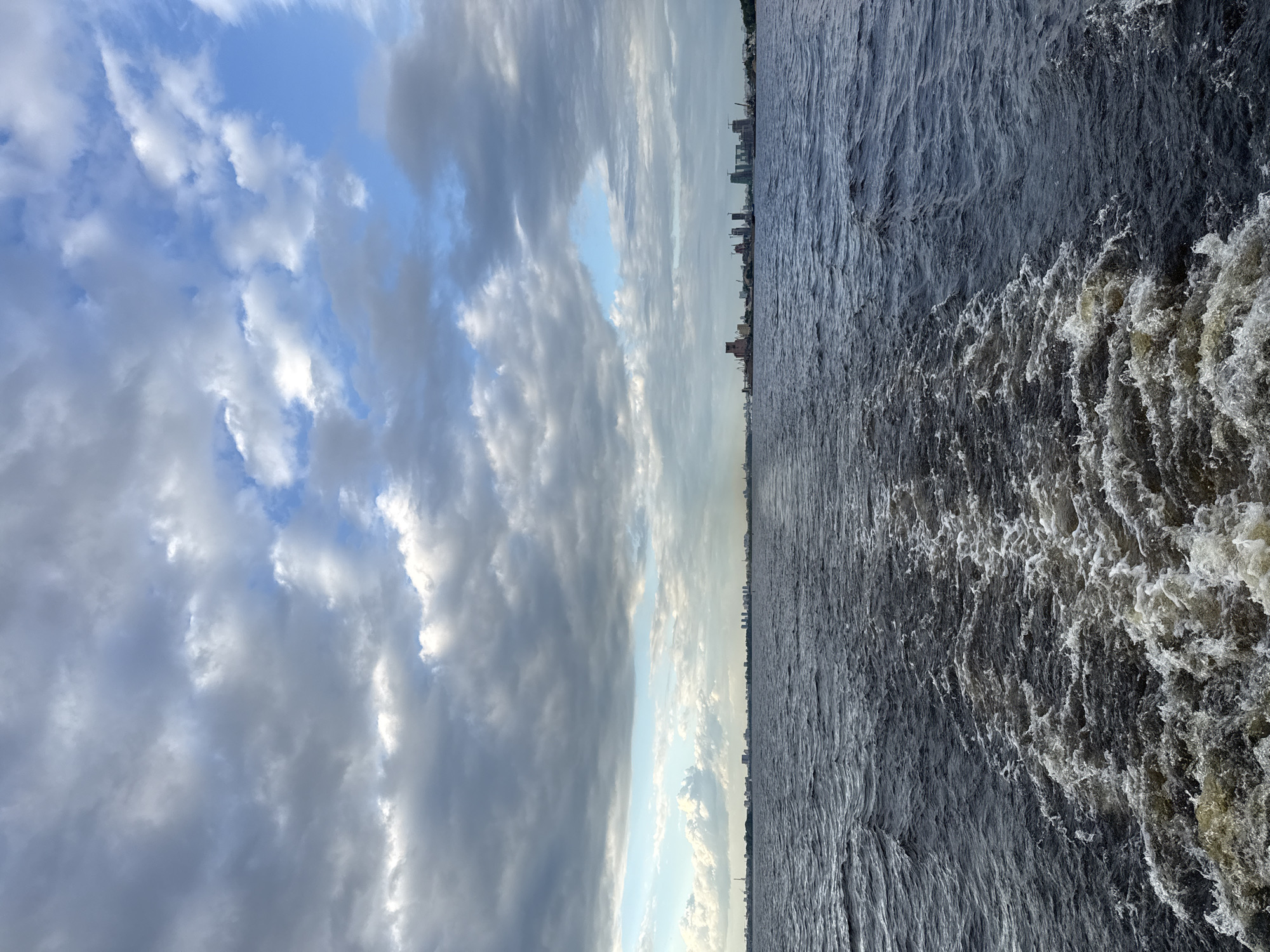 The image depicts a view from a boat moving away from the Kazan skyline, creating a wake in the dark, choppy water. The horizon is lined with buildings and structures, suggesting an urban environment. Above, a partly cloudy sky features a mix of dark gray and lighter white clouds, with patches of blue sky visible. The scene is captured from a low angle, looking back towards the city. There are no people or animals visible in the frame.