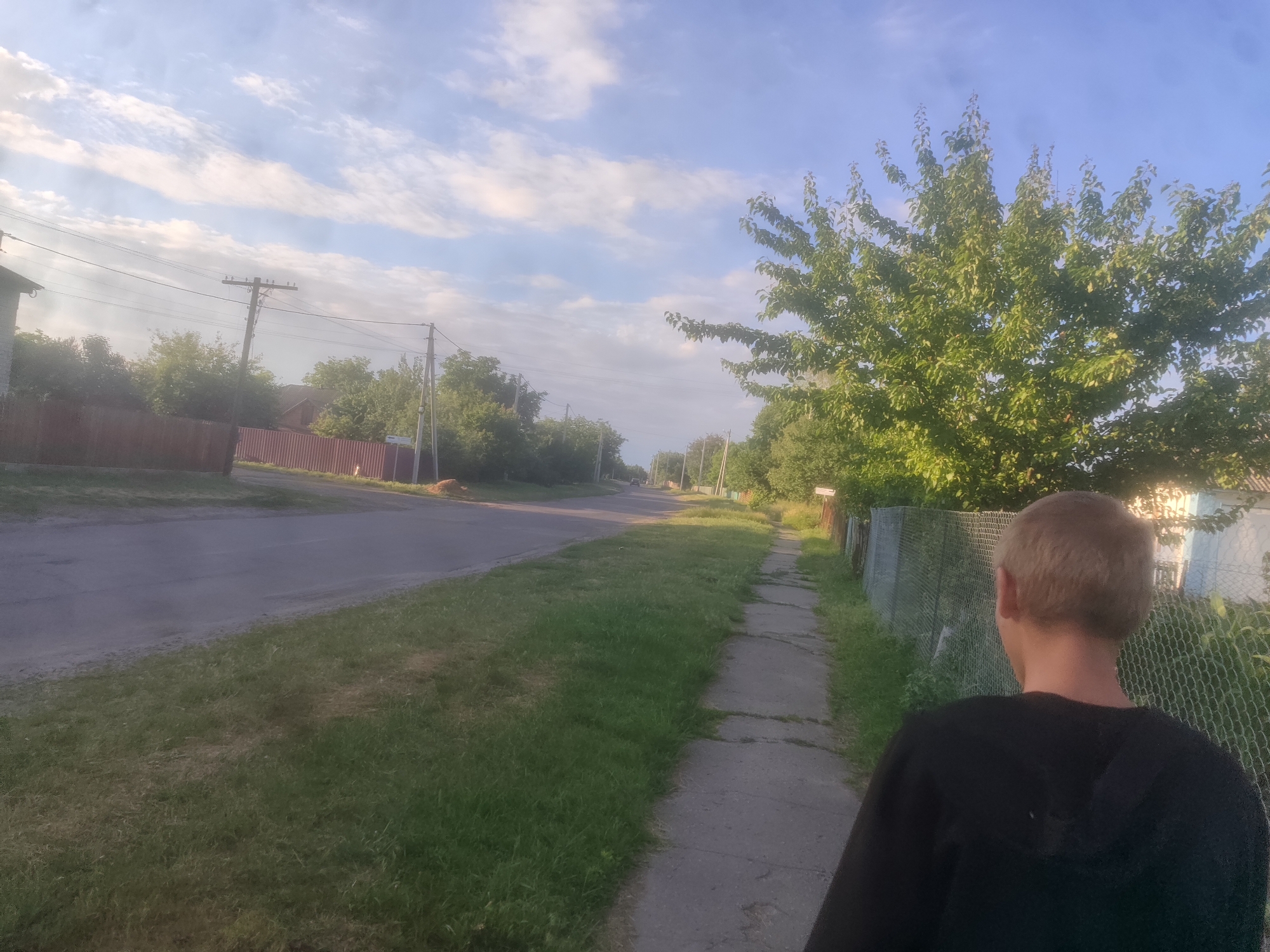 A young person with blond hair, wearing a black hooded sweatshirt, walks away from the camera on a cracked concrete sidewalk. The sidewalk is bordered by a chain-link fence on the right and a grassy area on the left. Beyond the grassy area is a paved road. In the background, trees and houses are visible under a sky with scattered clouds. Electrical poles and wires are also present along the road.