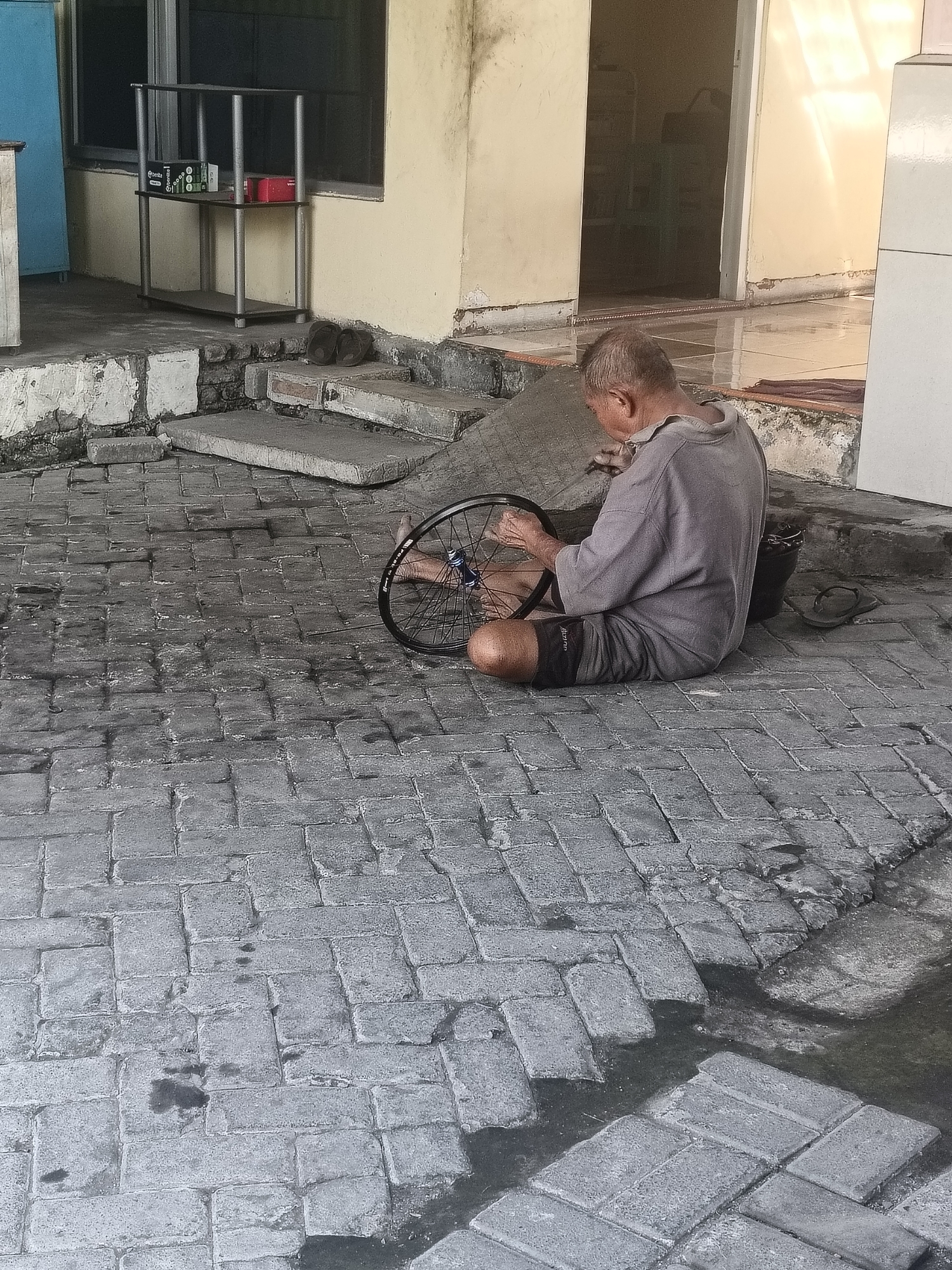 An elderly man, with thinning grey hair, is seated on the ground on a paved outdoor area. He is wearing a grey t-shirt and dark shorts. In front of him, on the ground, is a bicycle wheel which he is working on. To his right, on the ground, is a black bucket and a pair of dark flip-flops. Behind him, to his left, is a set of concrete steps leading up to a building with yellow walls and dark windows. A metal shelf unit with items on it is visible against the wall. The paved area appears to be made of rectangular bricks and some areas are wet. The scene is outdoors, with sunlight casting shadows. The location is identified as Gedangan, Indonesia.