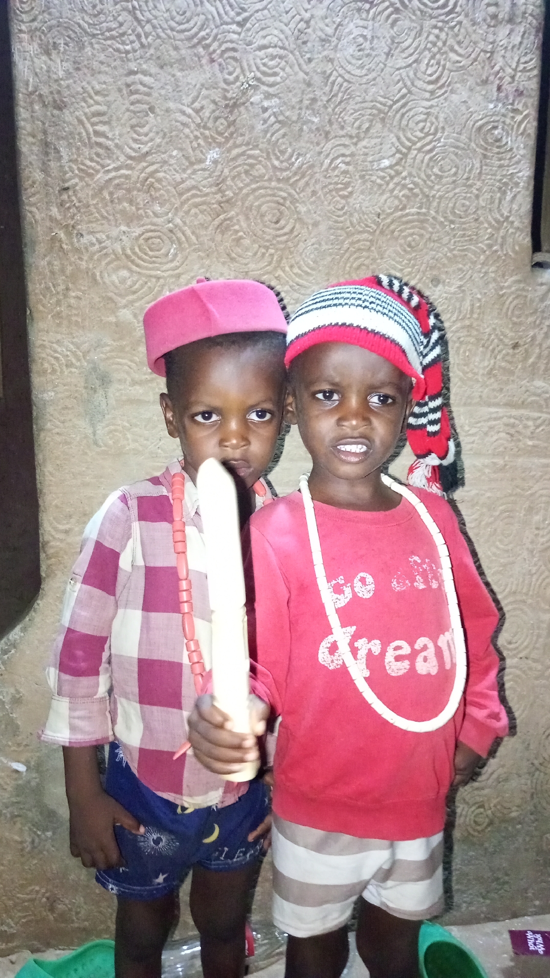 Two young children stand in front of a patterned wall. The child on the left wears a pink hat, a red and white checkered shirt, blue shorts with a moon and stars pattern, and a beaded necklace; this child holds a long, white object. The child on the right wears a red and white striped hat with a tail, a red sweatshirt with the word "dream" on it, white and gray striped shorts, and a white beaded necklace. They stand in a dimly lit room; the wall behind them features a textured, swirled pattern. The location is Onitsha, Nigeria.