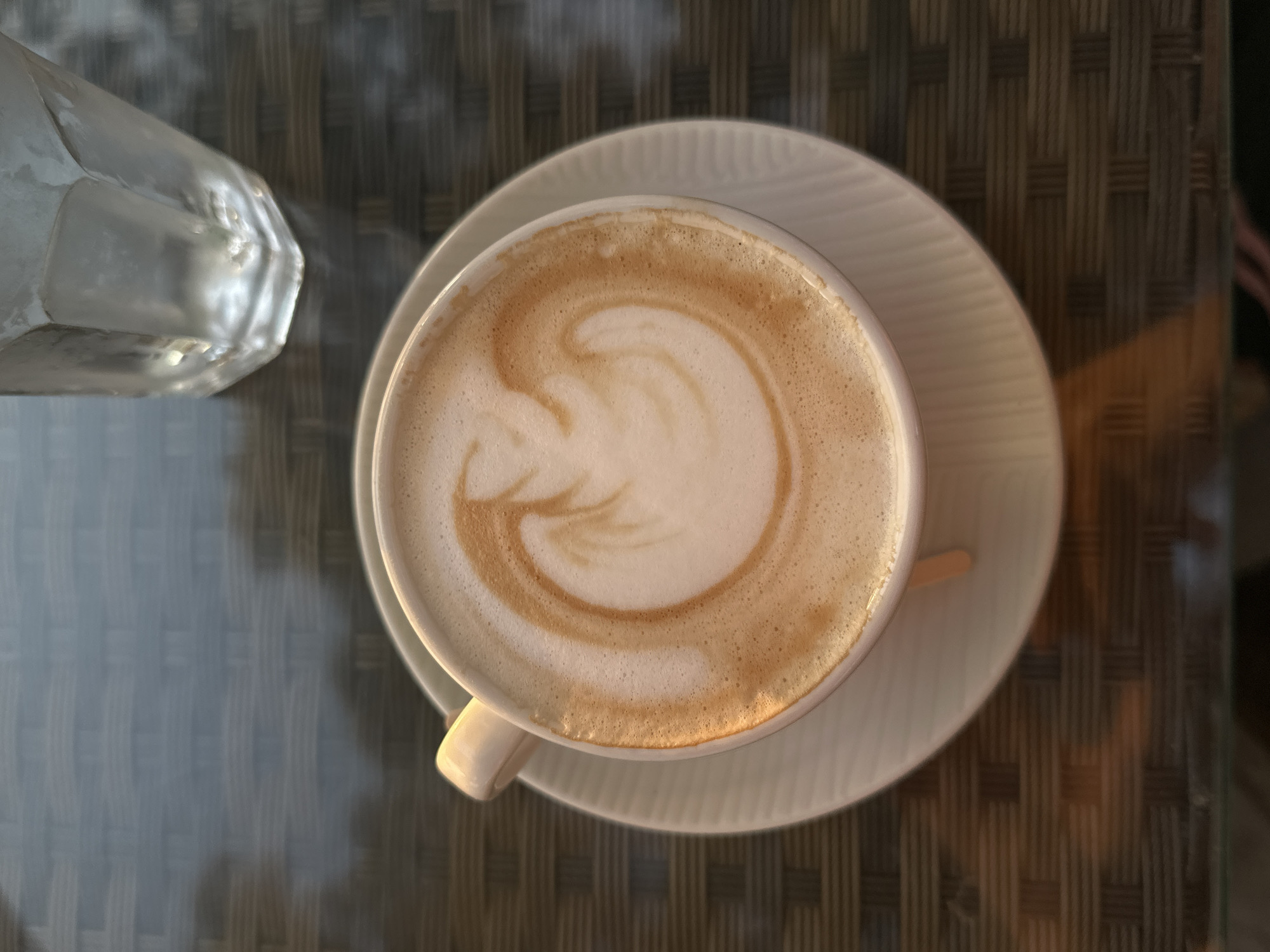 An overhead shot captures a cup of coffee and a partial view of a glass on a textured table. The central focus is a white ceramic cup filled with a light brown beverage, likely a latte or cappuccino, adorned with intricate latte art on its frothy surface, resembling a stylized swan or heart. The cup rests on a matching white saucer featuring subtle radial ridges.
To the upper left, a faceted clear glass, possibly containing water, is partially visible, reflecting the ambient light. The table surface beneath the items is dark and displays a distinct woven or checkered pattern, visible through what appears to be a transparent top, possibly glass.
The scene is illuminated by warm, directional light, suggesting either early morning or late afternoon sun. This lighting creates soft shadows and highlights, particularly on the glass and the rim of the coffee cup, contributing to a peaceful and inviting atmosphere. The image depicts a quiet moment, perhaps a coffee break at a cafe in Šušanj, Montenegro. No visible text is present.