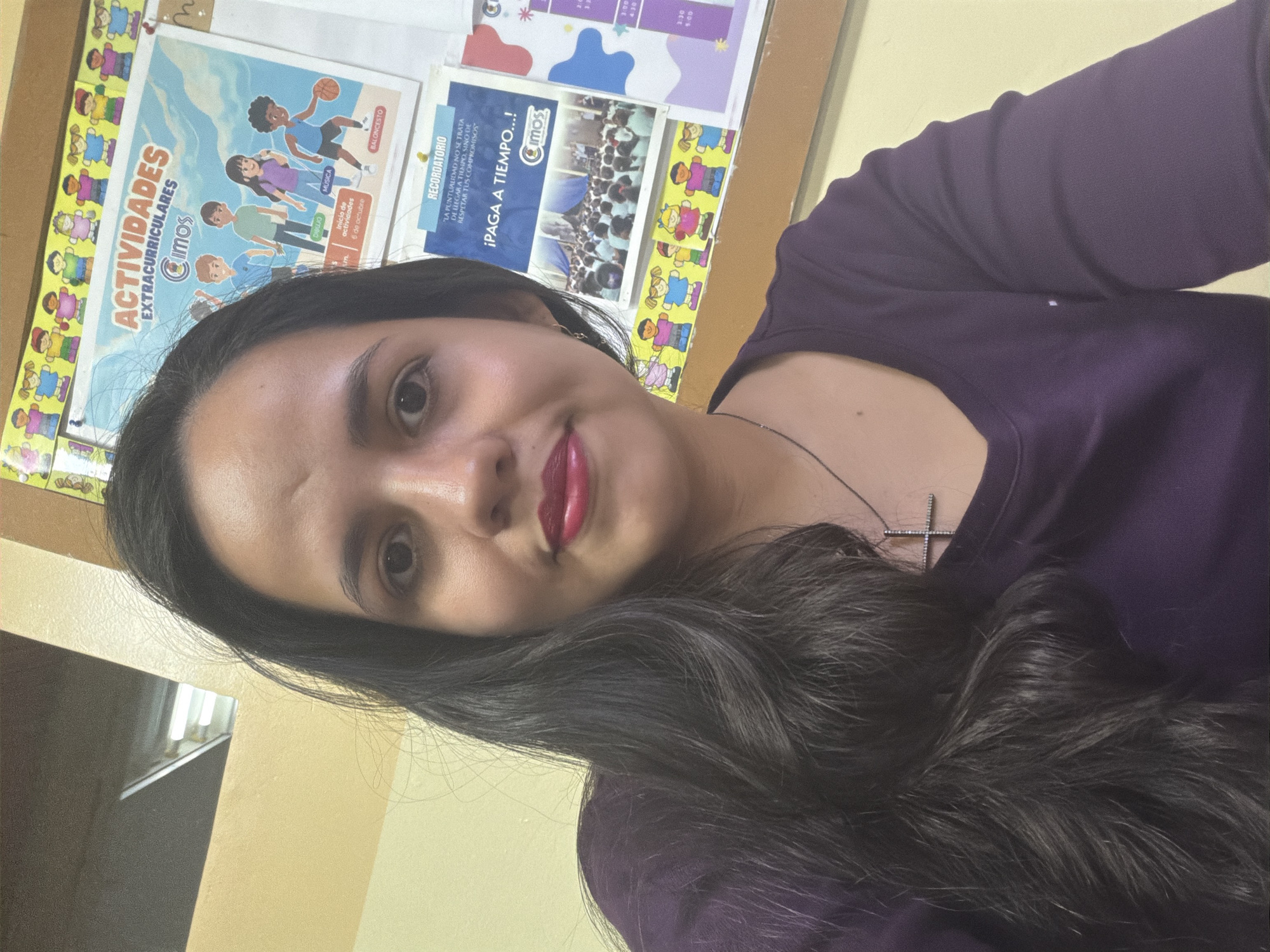 A woman with long, dark hair is posing for a selfie indoors. She is wearing a purple top and a necklace with a cross pendant. Behind her, a bulletin board displays colorful posters with the logo "Cimos," suggesting an educational or extracurricular context. One poster advertises "Actividades Extracurriculares," with images of children participating in music, basketball, and drawing. Another poster encourages timely payments. Based on the context provided by the "Cimos" logo, it can be inferred that this image was taken in Guayana City, Venezuela.