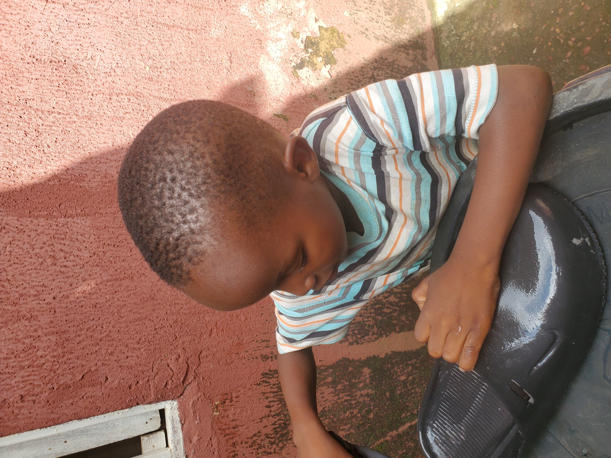 The image shows a young boy wearing a striped t-shirt, leaning over a large, black object that appears to be a bucket or container filled with water. His head is bowed, and he is looking down at his hands, which are resting on the edge of the object. The boy's expression is difficult to discern, but his posture suggests concentration or a moment of quiet activity. The surrounding environment is textured and appears to be outdoors, with a reddish-brown wall on the left and a mottled, greenish-brown ground on the right, possibly concrete with some moss or algae growth. Sunlight casts a shadow on the wall, indicating it is daytime. A sliver of a wooden structure is visible at the bottom left of the image. The overall scene suggests a simple, everyday moment, possibly involving a chore or play. The location context of Jos, Nigeria, points to a warm climate, consistent with the outdoor setting and the boy's attire.