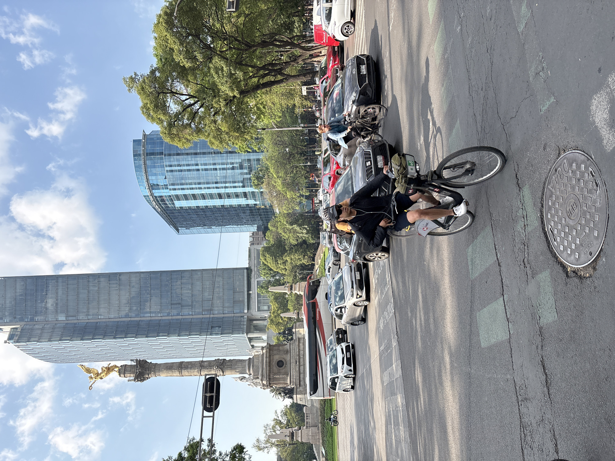 The image depicts a street in Mexico City, Mexico. In the background is the Ángel de la Independencia monument and modern glass skyscrapers. On the right side of the street, a line of cars is visible. Two people are riding bicycles in the bike lane. A manhole cover is visible on the right. The sky is blue with a few clouds. Trees line the street.