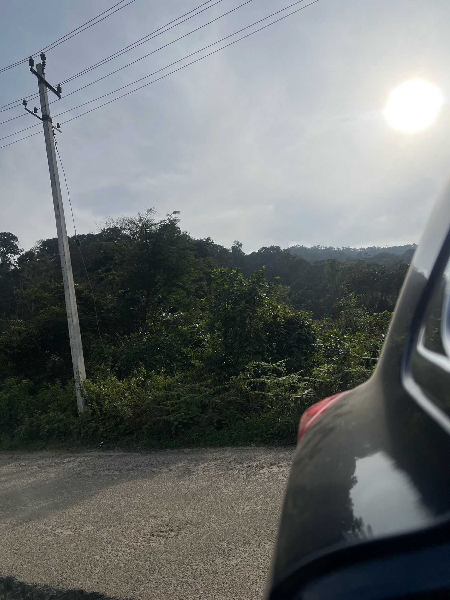 The image shows a road in the city of Hebbettageri, India. On the left side of the road, there is a utility pole with wires. The road is asphalt and appears to be in fair condition with some texture. Beyond the road, there is lush green vegetation covering a hillside. The vegetation consists of various trees and shrubs, creating a dense forest-like appearance. In the upper right portion of the image, the sun is visible, shining brightly in a hazy sky. The foreground of the image features a portion of a dark-colored vehicle, likely a car or motorcycle, its curved surface reflecting the sky and surrounding environment. The scene suggests an outdoor, natural setting along a road.