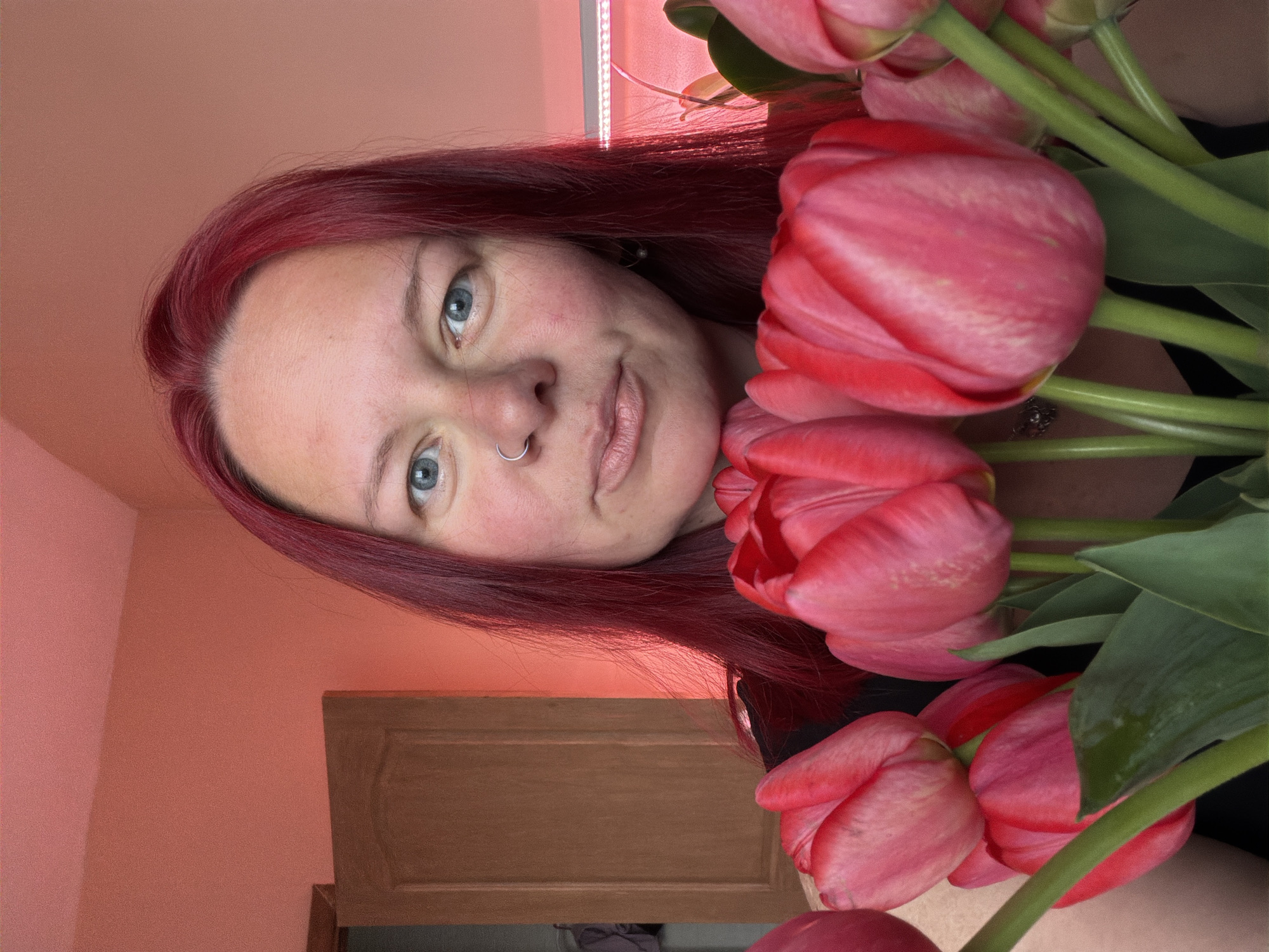 A fair-skinned woman with red hair poses with a bouquet of pink tulips. She has blue eyes and a silver nose ring. The background includes a pink wall, a wooden door, and a strip light. The location is unknown, as there is no evidence to indicate it is Chaikovsky, Russia.