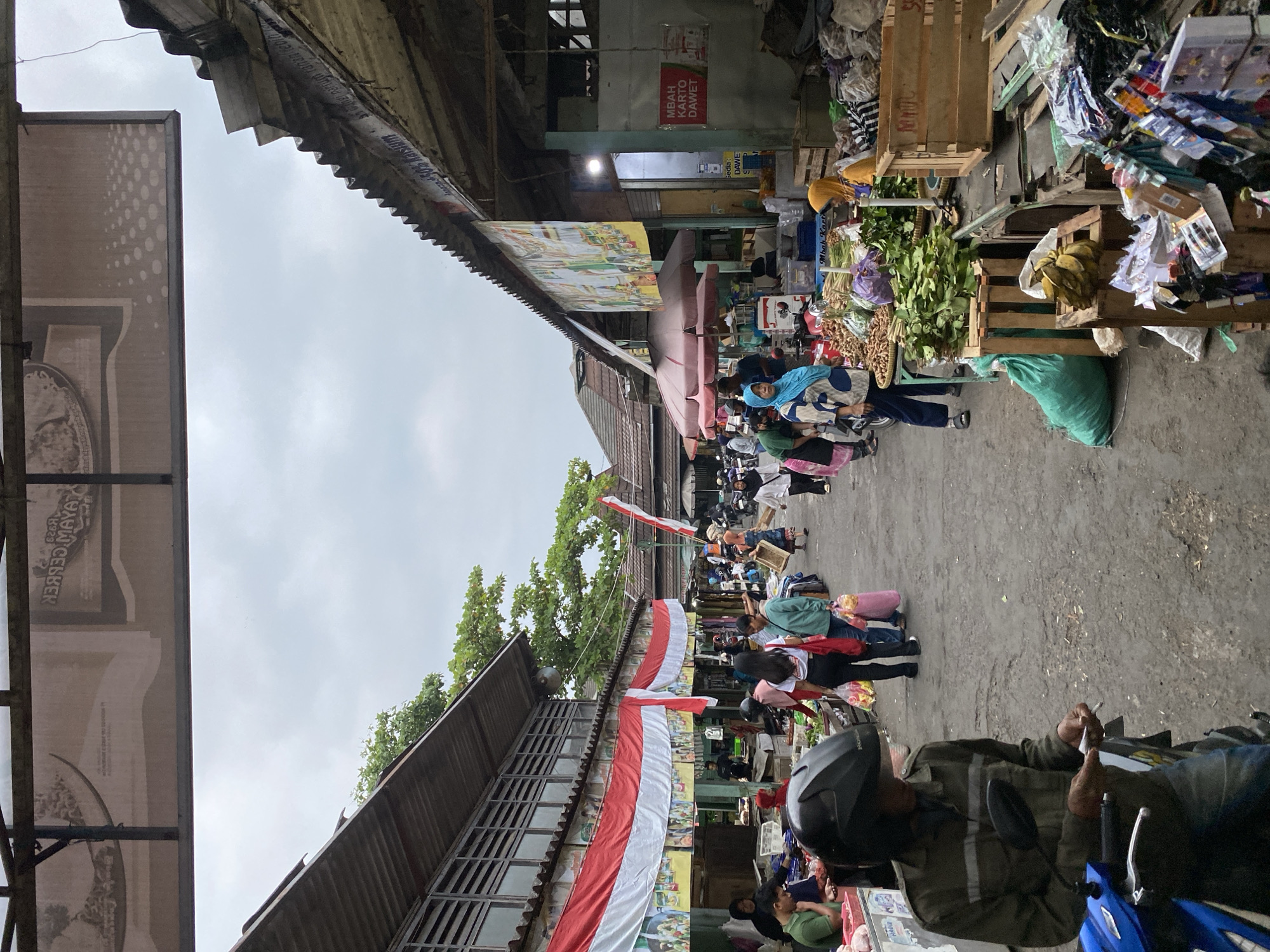 The image depicts a bustling outdoor market scene in Ambarketawang, Indonesia, under an overcast sky. The market appears to be a traditional one, with stalls lining a paved pathway. On the left, a long building with a corrugated metal roof and open-air windows features a prominent red and white Indonesian flag draped across its facade. People are seen browsing and shopping at various stalls.
In the foreground on the left, a person wearing a green jacket and a black helmet is seated on a blue motorcycle, holding a cigarette and what appears to be a lighter. The pathway is uneven and worn.
Further into the market, several individuals are visible. One person is wearing a light blue hijab and patterned clothing, standing near a stall laden with produce, including what look like bundles of leafy greens and turmeric. To their right, another person in a yellow hijab is tending to a stall with more produce and goods. Wooden crates are used for display and storage.
The stalls themselves offer a variety of items, including fresh produce, packaged goods, and possibly household items. Signs are visible on some of the stalls, some with text in Indonesian. The overall atmosphere suggests a lively commercial environment. The architecture of the market buildings, with their exposed beams and corrugated metal roofs, is typical of many traditional markets in the region. Trees with green foliage are visible in the background, suggesting the presence of vegetation within or surrounding the market area.