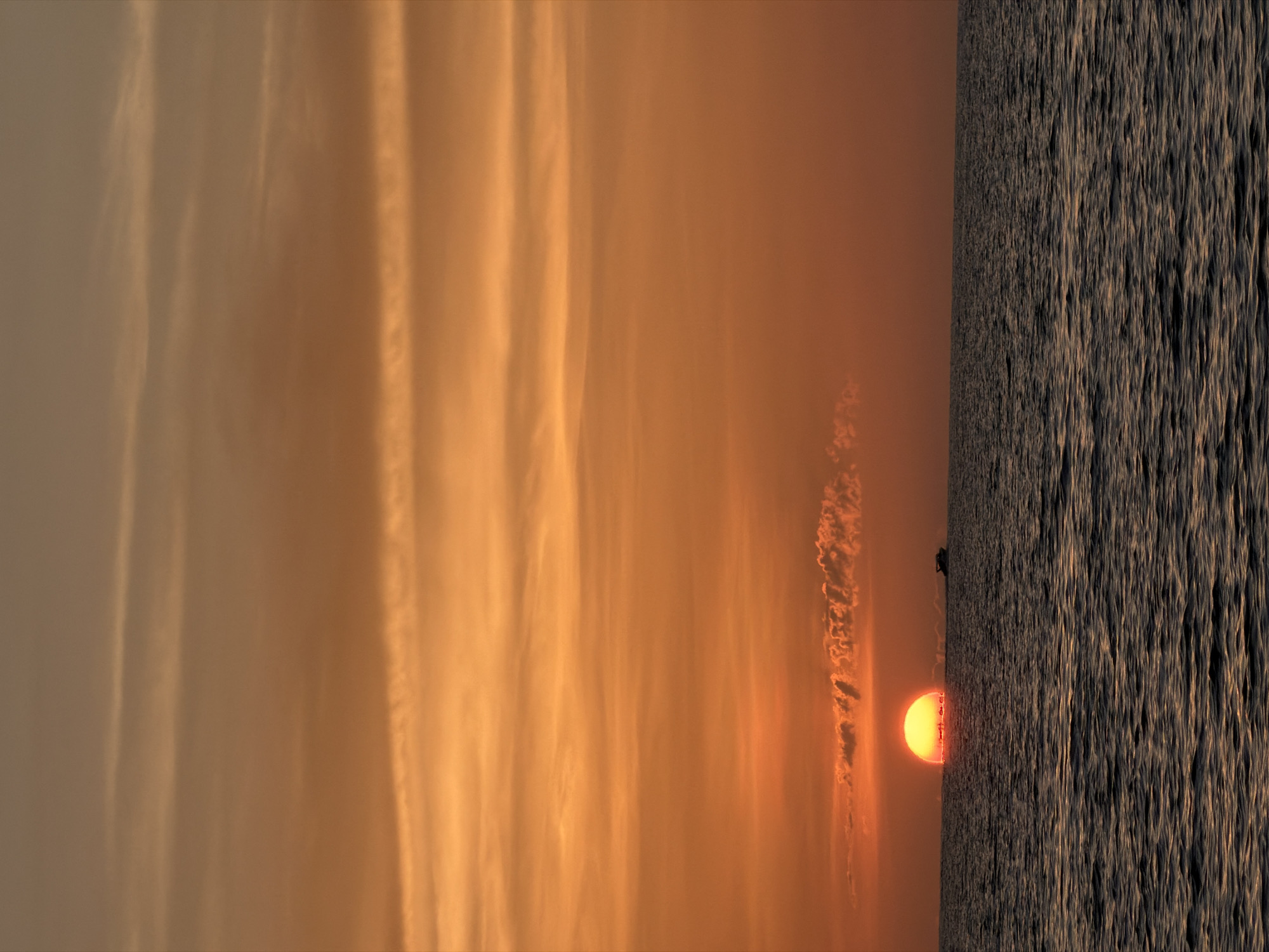 A seascape at sunset features a large, bright orange sun half-submerged on the horizon, casting a warm glow over the scene. The sky is filled with horizontal bands of orange and lighter hues, with thin, wispy clouds scattered across the upper half. The ocean, in the foreground, is a dark, rippling expanse reflecting the colors of the sky. A small, dark boat is visible to the right of the sun, moving across the water. The image captures the serene beauty of a sunset over the sea, likely from a coastal area of Ko Samui District, Thailand.