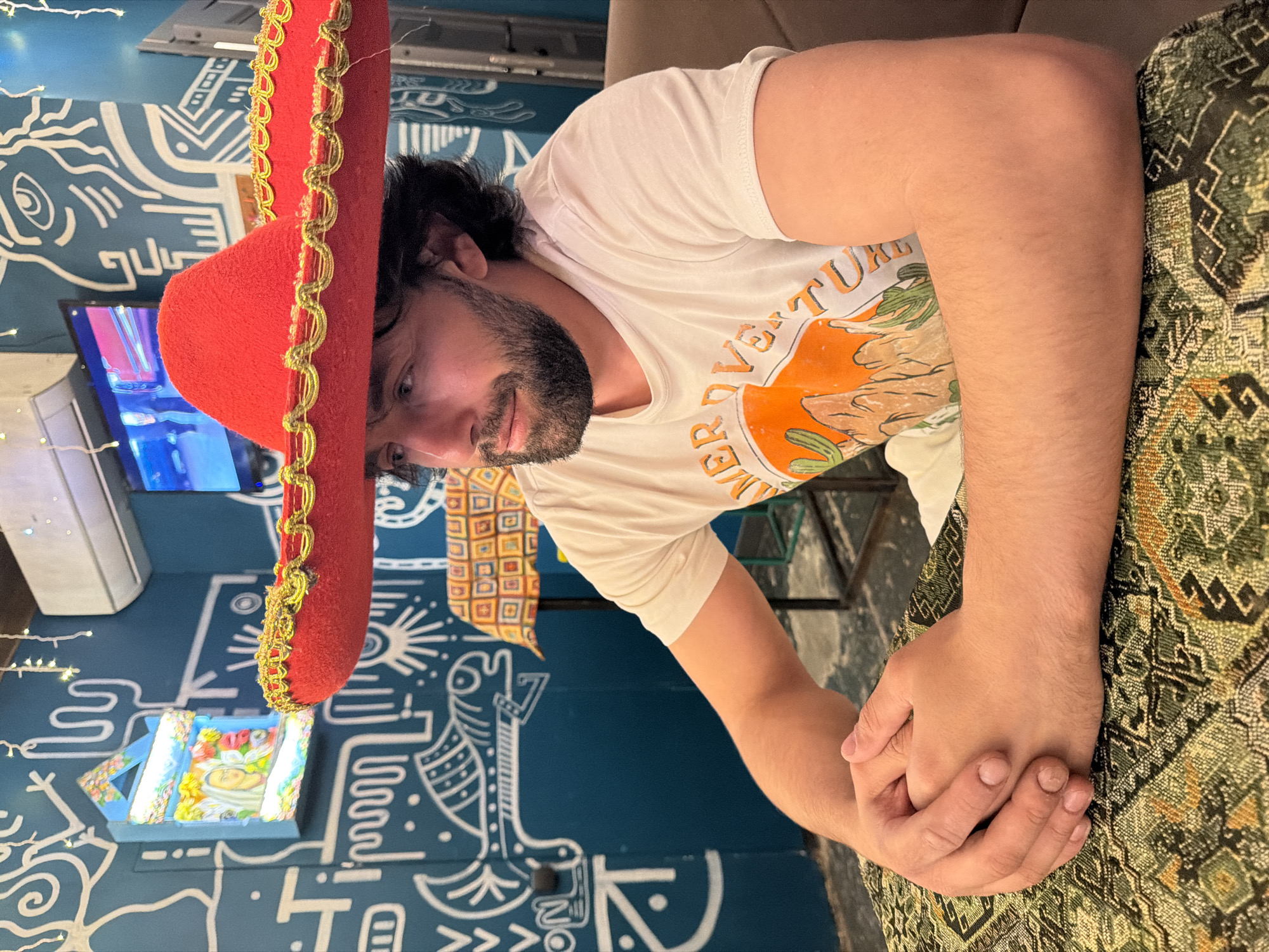 A person with dark hair and a beard is wearing a white t-shirt and a red sombrero with gold trim. They are seated indoors, with their hands clasped together on a table covered with a patterned fabric. Behind the person is a wall decorated with various white line drawings on a blue background. There's also a small, ornate object on the wall, a television and air conditioning unit. Based on the visual elements and available information, it can be inferred that this photo was taken in a themed restaurant, in the city of Krasnoyarsk, Russia.