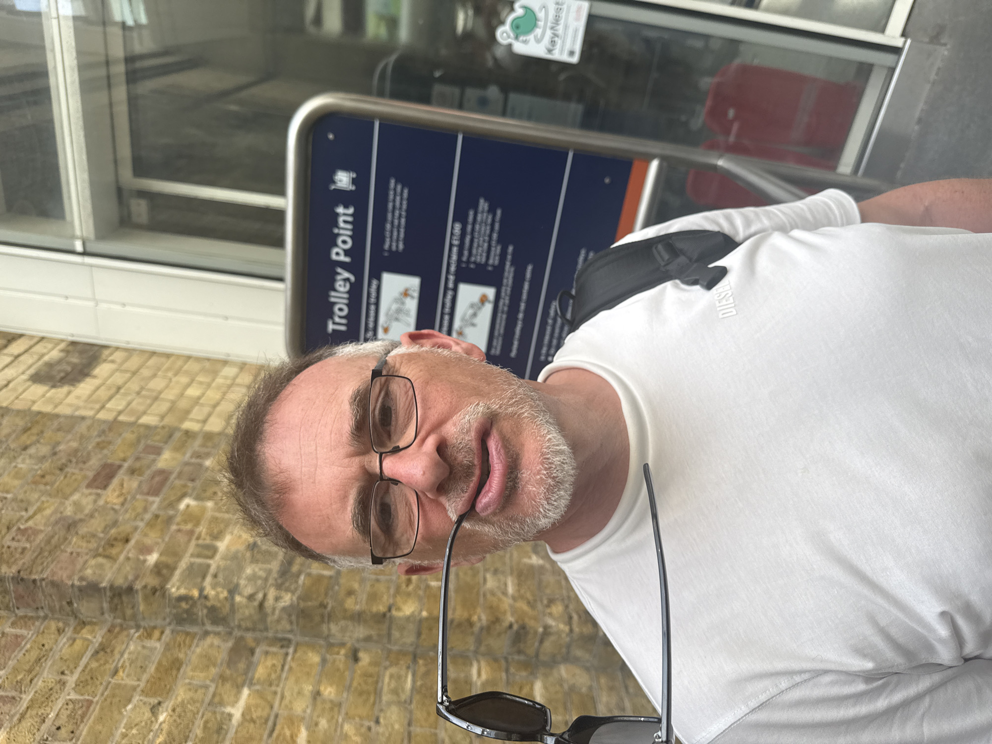 A middle-aged man wearing glasses and a white t-shirt stands beside a "Trolley Point" sign. He holds sunglasses in his mouth. He has graying hair and a salt-and-pepper beard. The background includes a brick walkway, a glass-fronted building, and a KeyNest sign. The location is likely a transportation hub where trolleys are available. The city is Unknown, Unknown.
