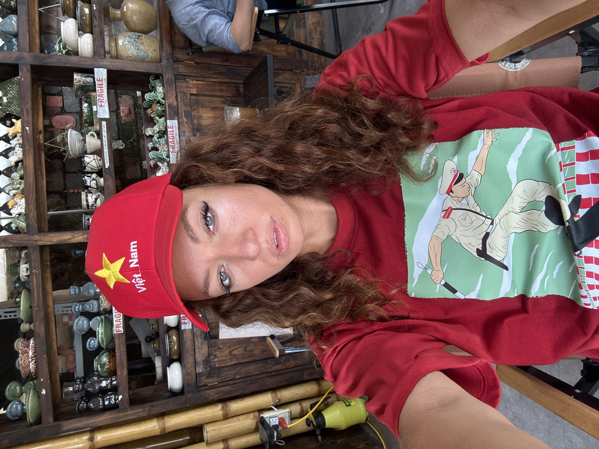 A selfie shows a woman wearing a red baseball cap with a gold star and the words "Việt Nam" in yellow lettering and a red, long-sleeved shirt with a graphic of a Vietnamese soldier on a green background, likely taken in a shop or cafe in Hà Nội, Vietnam. She has curly brown hair and fair skin. Behind her is a wooden shelf holding pottery and other decorative items, some labeled "fragile". A power outlet and a piece of equipment are seen on a bamboo structure to the left of the frame. The lighting is bright, suggesting it's daytime. Another person is partially visible in the upper left corner.