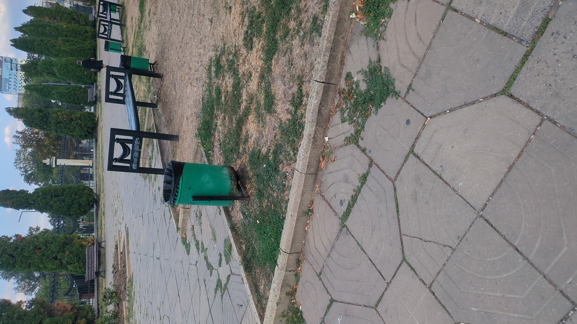 The image depicts a public park area in Chișinău, Moldova, characterized by paved pathways, grassy areas, and cypress trees. In the foreground, a green trash can is visible on a patch of grass next to a paved area. Further back, several black park benches with wooden slats are arranged along the pathways. A woman in a dark dress is walking away from the camera in the middle ground, towards the right side of the image. The background shows more trees, a black metal fence, and a modern building with glass windows. The overall scene suggests a serene, urban park environment.