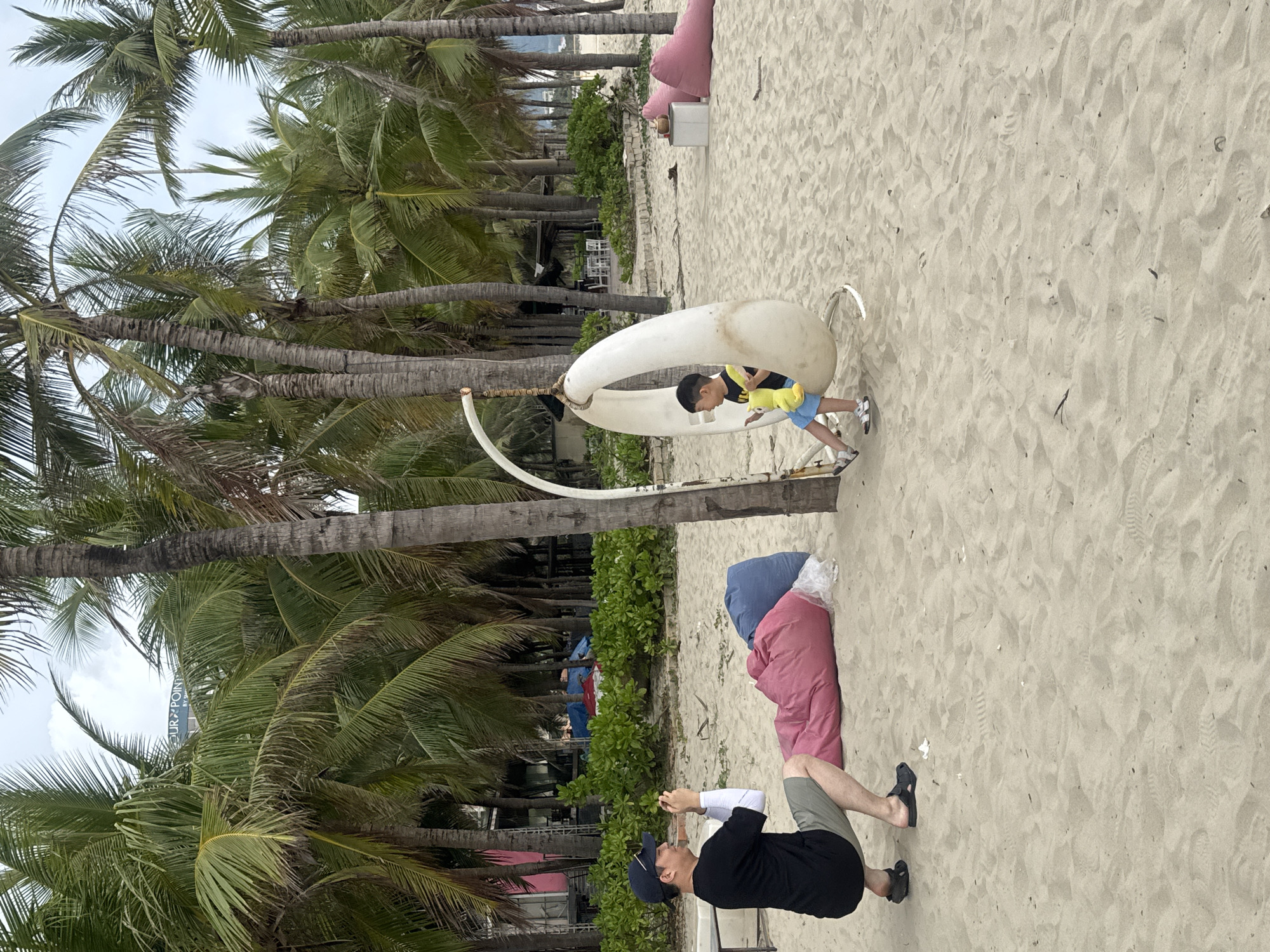 The image depicts a beach scene in Đà Nẵng, Vietnam. A man wearing a black shirt, khaki shorts, and a baseball cap is crouching, possibly taking a photo with a smartphone. A young boy in a yellow shirt and blue shorts stands inside a white hanging chair. The beach has several palm trees, beanbags, and some greenery. The sand is light and shows footprints. The sky is overcast. A building with the words "TOUR POINT BY" can be seen through the trees.