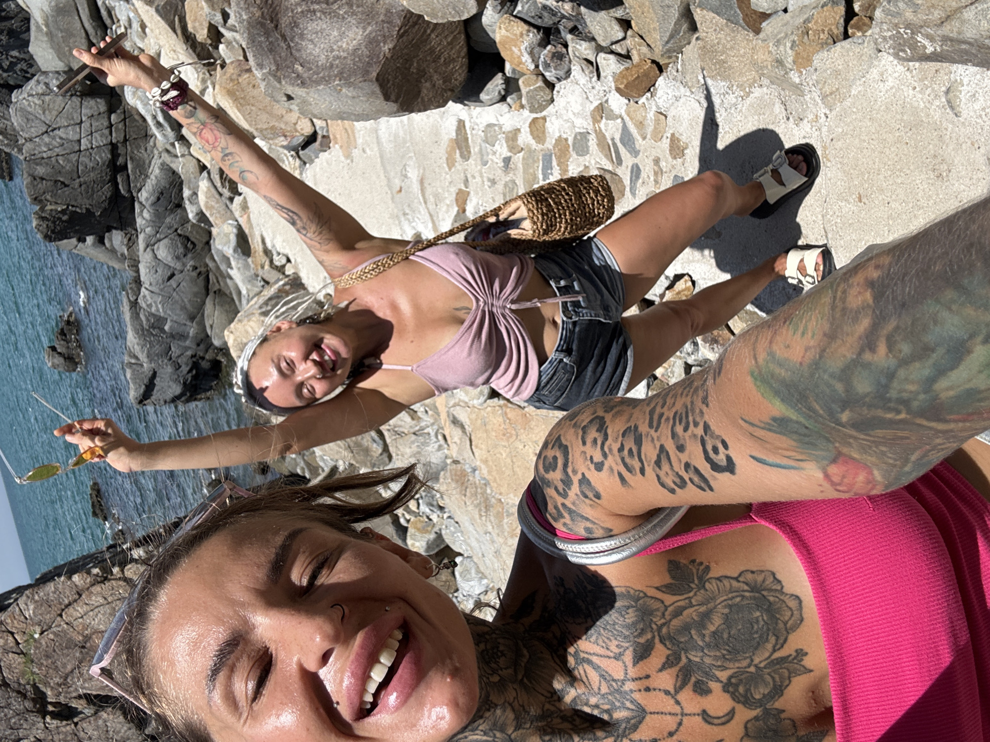 Two women are standing on a rocky, coastal path in Vĩnh Lương, Vietnam. The woman in the foreground is taking a selfie, smiling broadly with her eyes closed. She has a full sleeve of tattoos on her left arm and chest, wearing a bright pink tank top. The woman in the background, slightly behind and to the right, is also smiling and holding up sunglasses in her right hand. She has lighter hair styled in braids and is wearing a pink ruched crop top and denim shorts. She has a woven bag slung over her shoulder. The background shows the blue ocean and rocky terrain characteristic of the Vĩnh Lương coastline. The path they are standing on appears to be paved with stones and concrete.