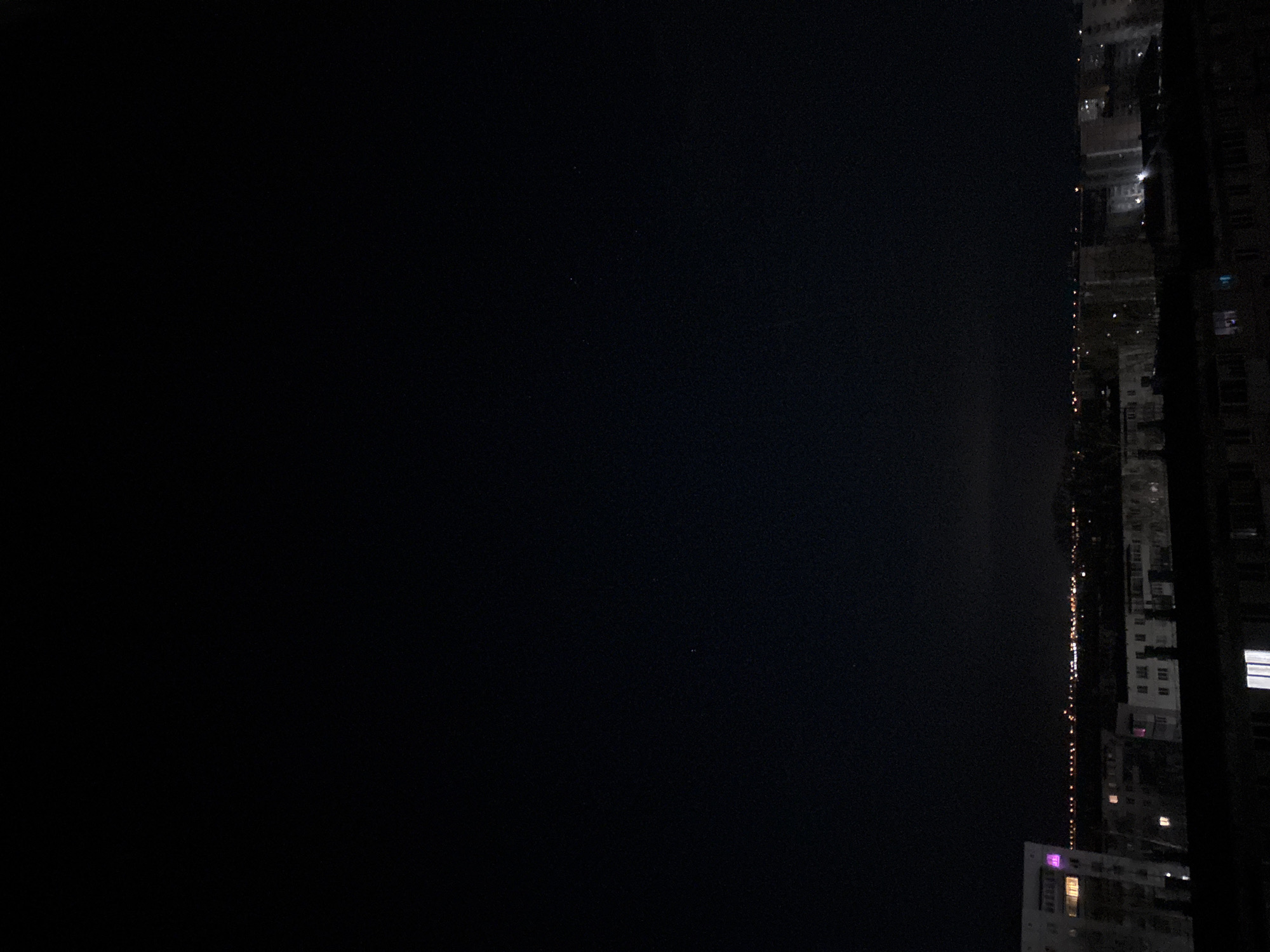 An eye-level shot shows a dark night sky beside a high-rise building. The night sky fills most of the frame, with a handful of barely discernible stars. To the right of the frame, the building stands tall, with a few windows illuminated. A line of lights hints at a street or road adjacent to the structure. The scene appears to be captured from an elevated position, possibly from another high-rise or a high floor within the same building. There is no evidence that this is in Chaikovsky, Russia. There are no people visible.