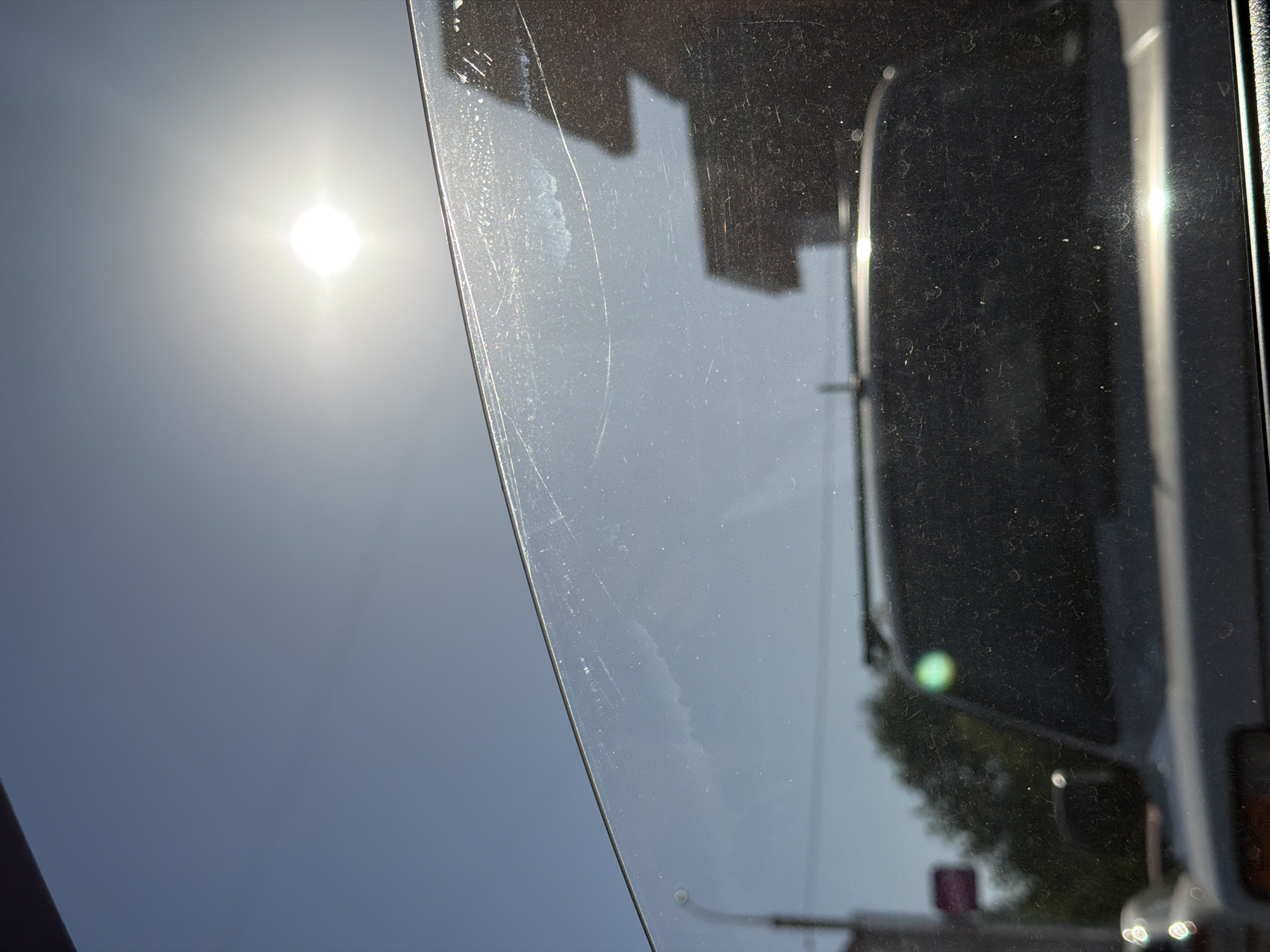 The image is taken from inside a vehicle, looking out through a slightly dirty and scratched window. Outside the window, the sun is shining brightly in a clear blue sky. Reflections of buildings and a vehicle, potentially a bus or truck, are visible in the window. The presence of foliage suggests an urban area with trees. While the location cannot be definitively confirmed as Izhevsk, Russia, the buildings and general appearance are consistent with an urban environment.
