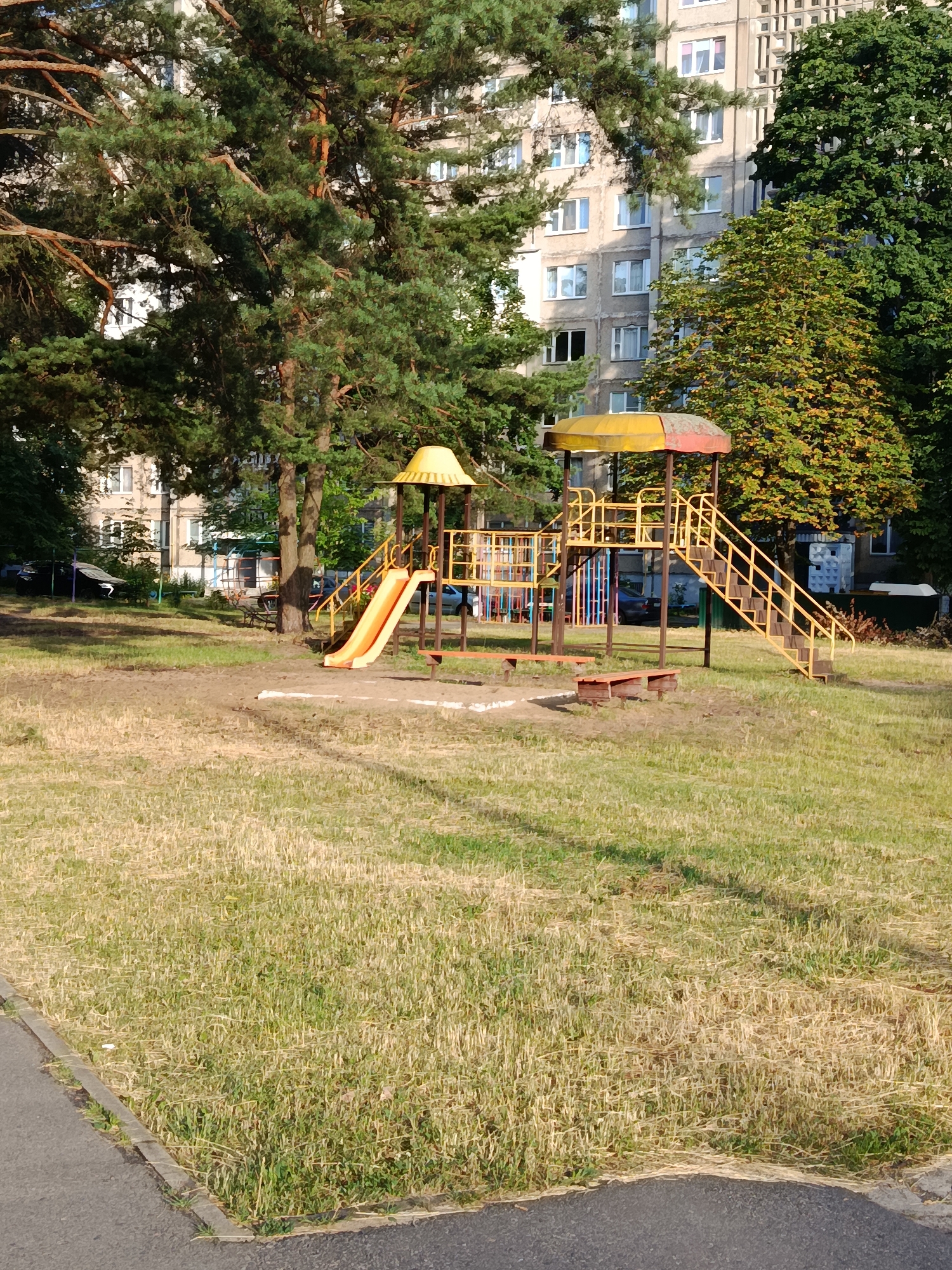 The image shows a playground in the city of Svietlahorsk, Belarus. The playground features a yellow metal structure with a slide and stairs. There are also two wooden benches. The playground is surrounded by grass and trees, and a large apartment building is visible in the background. The sun is shining, casting shadows on the ground. The grass is dry and yellow in some areas, suggesting it might be summer. There are no people or animals visible in the image. The scene appears to be calm and quiet.