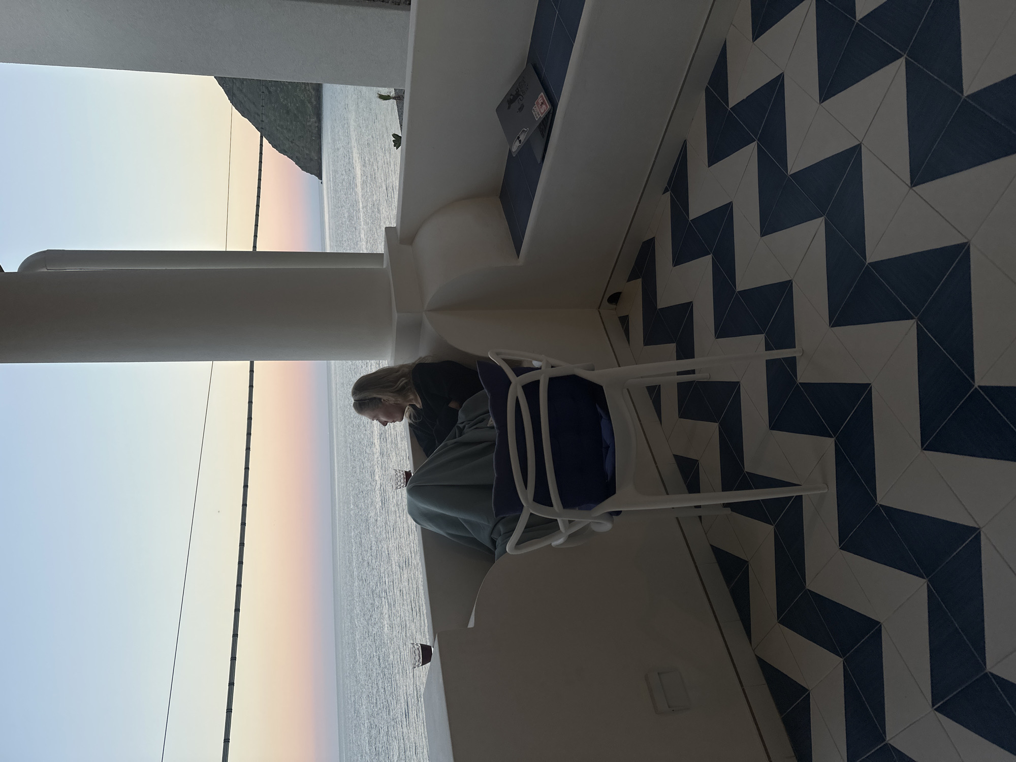 On a balcony overlooking the sea, a woman with blonde hair sits on a white chair, appearing to be working. A laptop rests on a ledge near a bench with dark blue accents. A throw and blue garment are draped on the chair. The balcony features white columns and a tiled floor with a blue and white geometric pattern. In the background, the sky transitions from blue to pink above the sea, and a landmass is visible on the horizon. The image depicts an outdoor setting, with natural light and elements that suggest the location is Lipari, Italy.