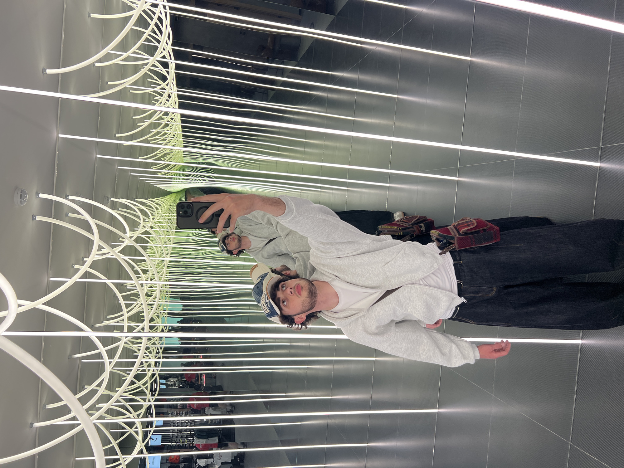 A male individual takes a selfie in Yerevan, Armenia. He is dressed in a white t-shirt, a light grey hoodie, dark jeans, and a blue and white hat. The setting appears to be inside a building, characterized by walls lined with parallel, illuminated white lines and curved light fixtures on the ceiling. The reflective surfaces create an illusion of a long, illuminated tunnel.