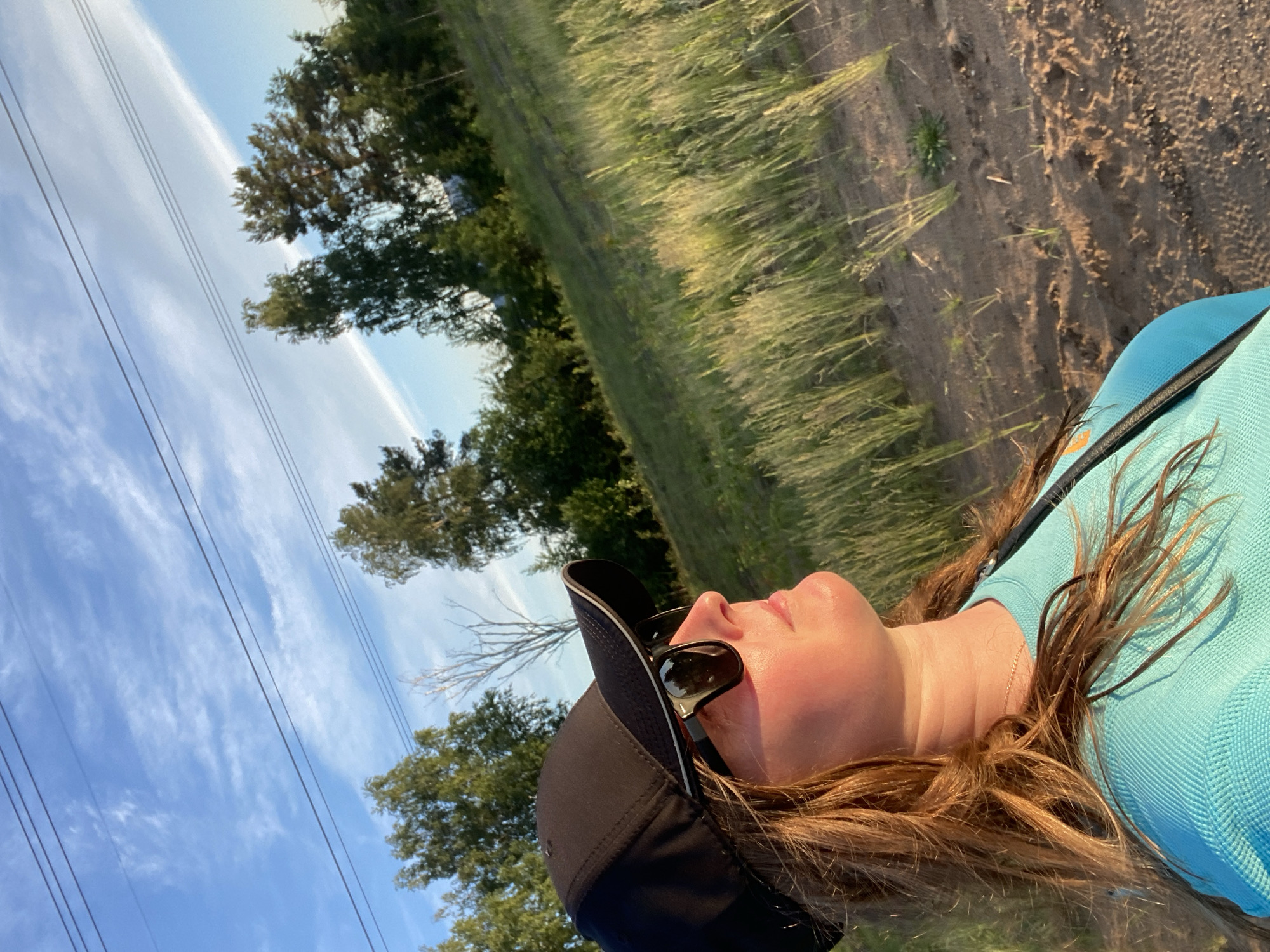 The image captures an outdoor scene in what appears to be late afternoon or early evening, oriented diagonally with the horizon line slanted. The primary subject is a person, likely female, positioned in the lower right foreground. She is seen from the chest up, looking upward and slightly to the right, with a relaxed expression. She has long, light brown hair, fair skin, and wears a light blue textured top, a black baseball cap, and black sunglasses resting on the cap. A thin gold chain is visible around her neck.
The background features a rural or semi-rural landscape under a bright blue sky with scattered white clouds. Several power lines stretch across the sky from left to right. To the left, a line of green-leafed trees stands against the sky. The midground and right side of the image show tall, green and golden-yellow grass, suggesting a field or roadside. Adjacent to the grass is a dirt path or track, showing variations in texture and some small green plants. The lighting casts warm tones on the grass and the person's hair, indicating low sunlight. No specific activity is depicted beyond being outdoors and observing the surroundings. There is no visible text in the image. The location context is Noviy, Russia.