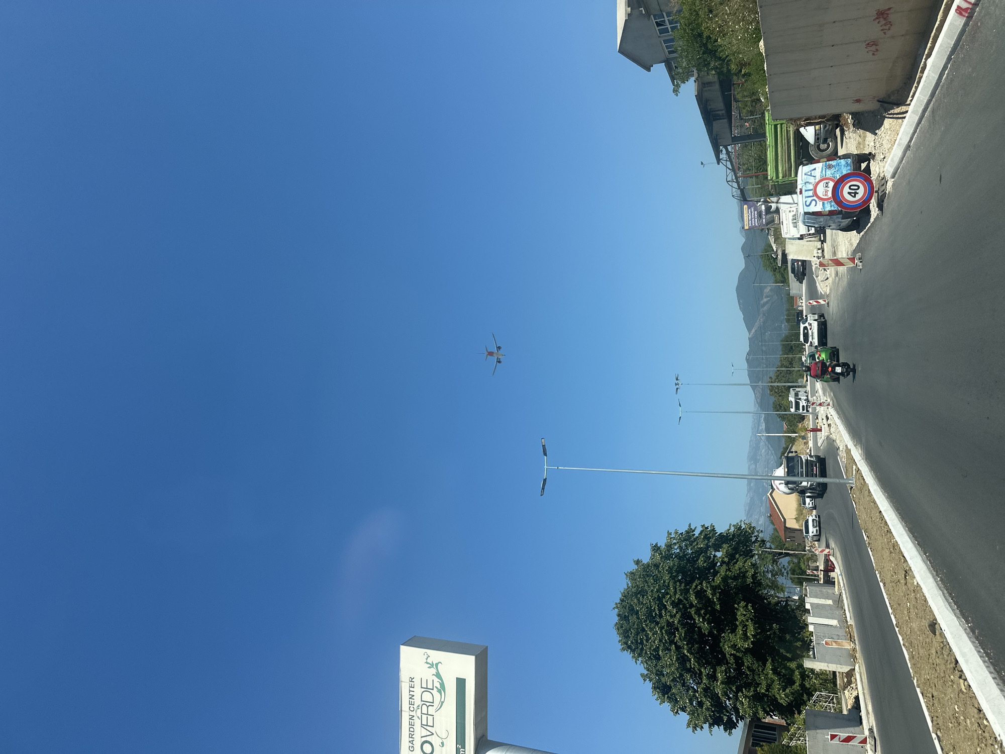 In Radanovići, Montenegro, under a clear blue sky, a road construction scene unfolds. The asphalt road, newly paved and bordered by freshly laid curbs and dirt, curves through the frame. On the left, a sign for "GARDEN CENTER OVERDE" stands tall. A large, lush green tree dominates the left mid-ground, with mountainous terrain visible in the far distance.
Several vehicles navigate the road: a motorcycle with a rider in red, a green car, a white sedan, and a white truck carrying a large tank. Further down the road, a green dump truck and a white van with the word "SUZA" and traffic signs on its side are parked. A circular speed limit sign indicating "40" is prominently displayed.
In the upper portion of the image, an airplane is captured in mid-flight against the vast blue sky. The scene suggests a day of ongoing activity, possibly related to development or transportation in the area. The presence of construction materials and workers' barriers indicates an active work zone.