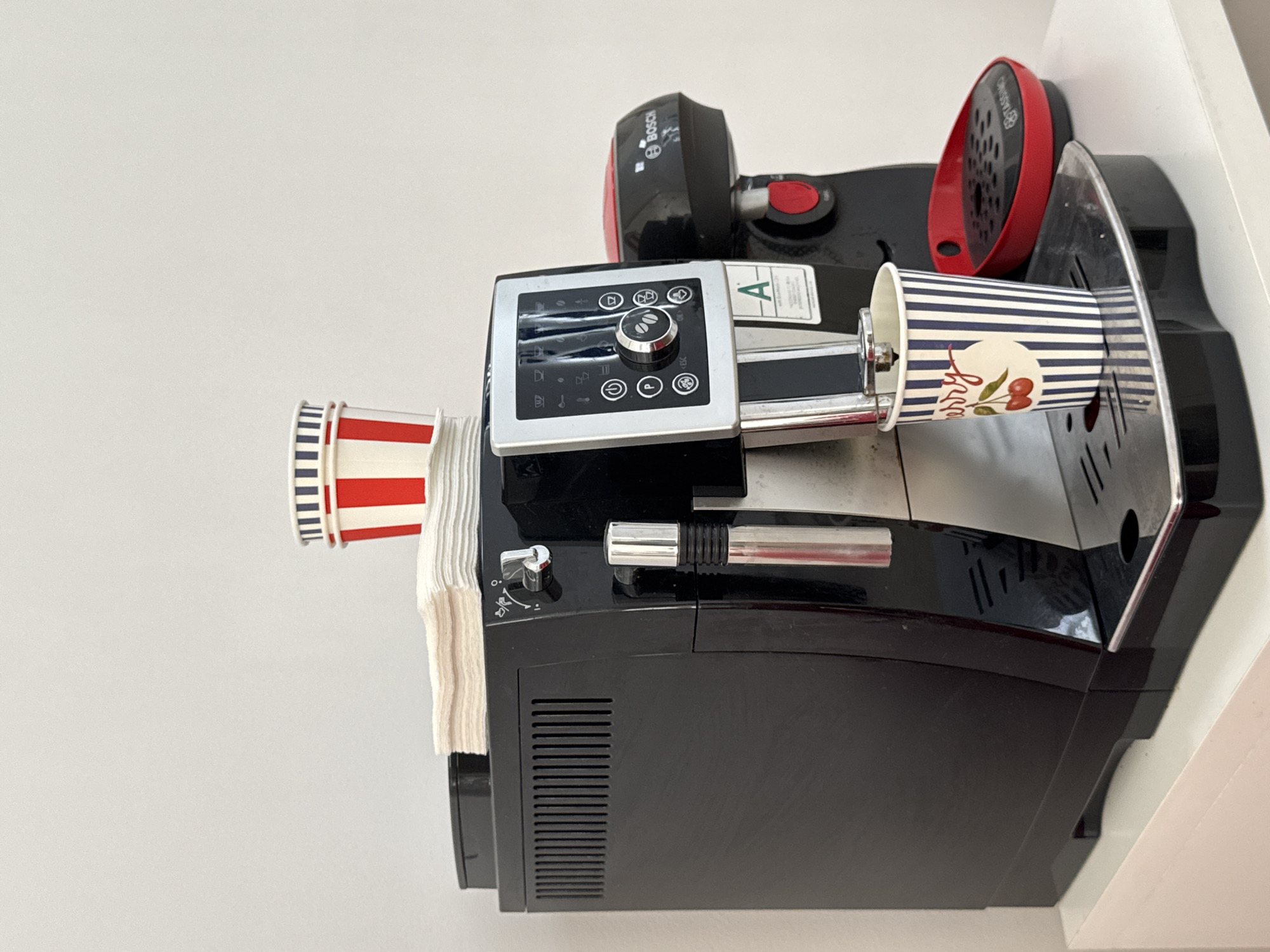 The image shows a close-up, eye-level, indoor shot of a black automatic coffee machine positioned on a white surface. On top of the machine, there are two red and white striped paper cups stacked on a pile of white napkins. To the right of the coffee machine, a smaller red and black Bosch appliance is visible. In front of the coffee machine, a single blue and white striped paper cup with a cherry design is placed on the drip tray. The background is a plain, light-colored wall. The scene is static, with no people or animals present. The lighting is soft and even, suggesting an indoor setting without direct sunlight. The image lacks any visual cues indicating its location in Yeysk, Russia.
