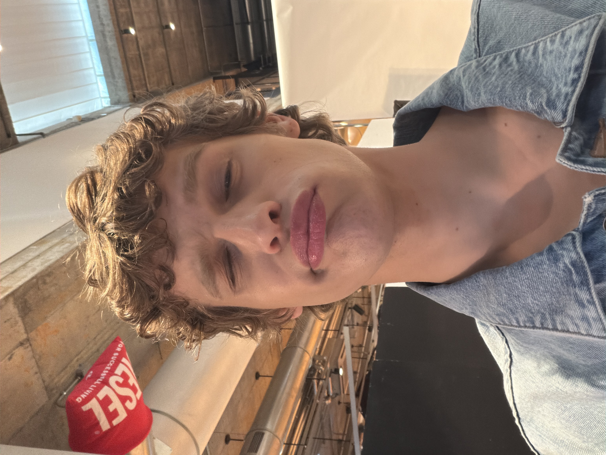 A medium shot captures a person with light skin and curly brown hair, donning a light blue denim jacket. The person is positioned at an unusual angle, tilting to the left, giving the impression of lying down. Their face is partially obscured by the angle, with one eye appearing closed or squinting.
The backdrop is an urban, industrial-looking setting, possibly inside a building. Visible architectural elements include metal pipes running along the ceiling and a window with blinds. A red, triangular object with the word "DIESEL" in white lettering is situated in the lower left corner.