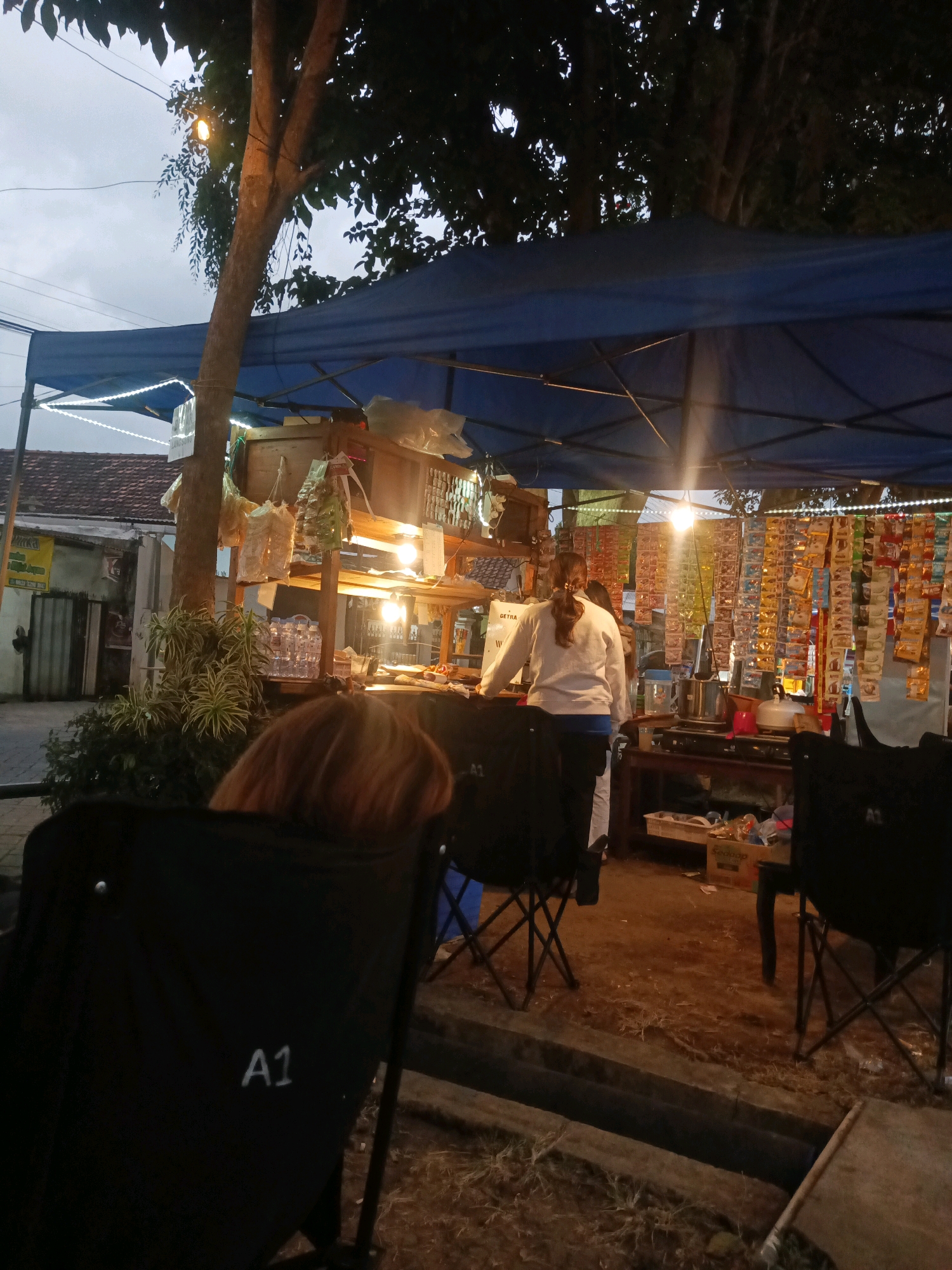 The image depicts an outdoor food stall at dusk, likely in Tulungagung, Indonesia. The stall is covered by a blue tarp, illuminated by string lights and a bright overhead bulb. Shelves behind the counter are stocked with various packaged goods and bottled water. A woman in a white sweatshirt and dark pants stands behind the counter, appearing to serve a customer. In the foreground, two black folding chairs are visible, one with an "A1" marking. The ground is dirt and scattered with debris. The overall atmosphere suggests a casual, local eatery.