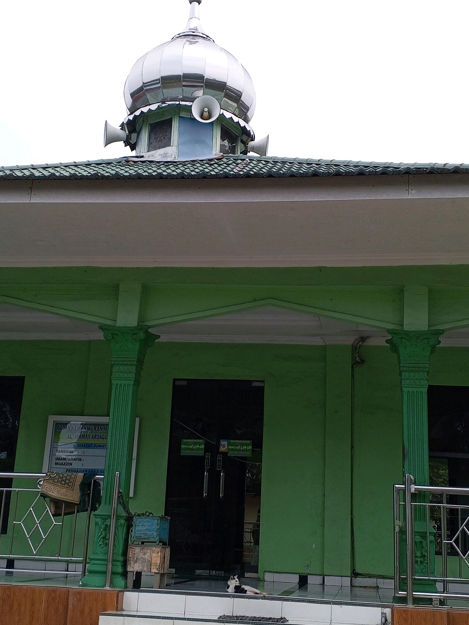The image shows the exterior of a mosque with a green facade and a silver dome. The dome has speakers attached to its base. The mosque has a covered entryway supported by green, fluted columns with decorative capitals. A sign is visible on the left side of the entryway, along with a metal railing. In front of the mosque, there are steps leading up to the entrance. A cat is lying on the tiled floor in front of the entrance. The sky in the background is overcast. The sign indicates that the mosque is located in Deli Tua Timur, Indonesia. The sign is in Indonesian and contains information about the mosque's administration, prayer times, and religious officials. Specifically, it lists the mosque as "BADAN KEMAKMURAN MASJID AL-HIKMAH ARDAKU", which translates to "Masjid Al-Hikmah Ardaki Prosperity Board". It also mentions "SHALAT JUMAT" (Friday Prayer) and lists roles like "IMAM/KHATIB" (Imam/Preacher) and "MUAZZIN" (Caller to prayer). A section for "PENGAJIAN AHAD SU" (Sunday Study) is also visible.