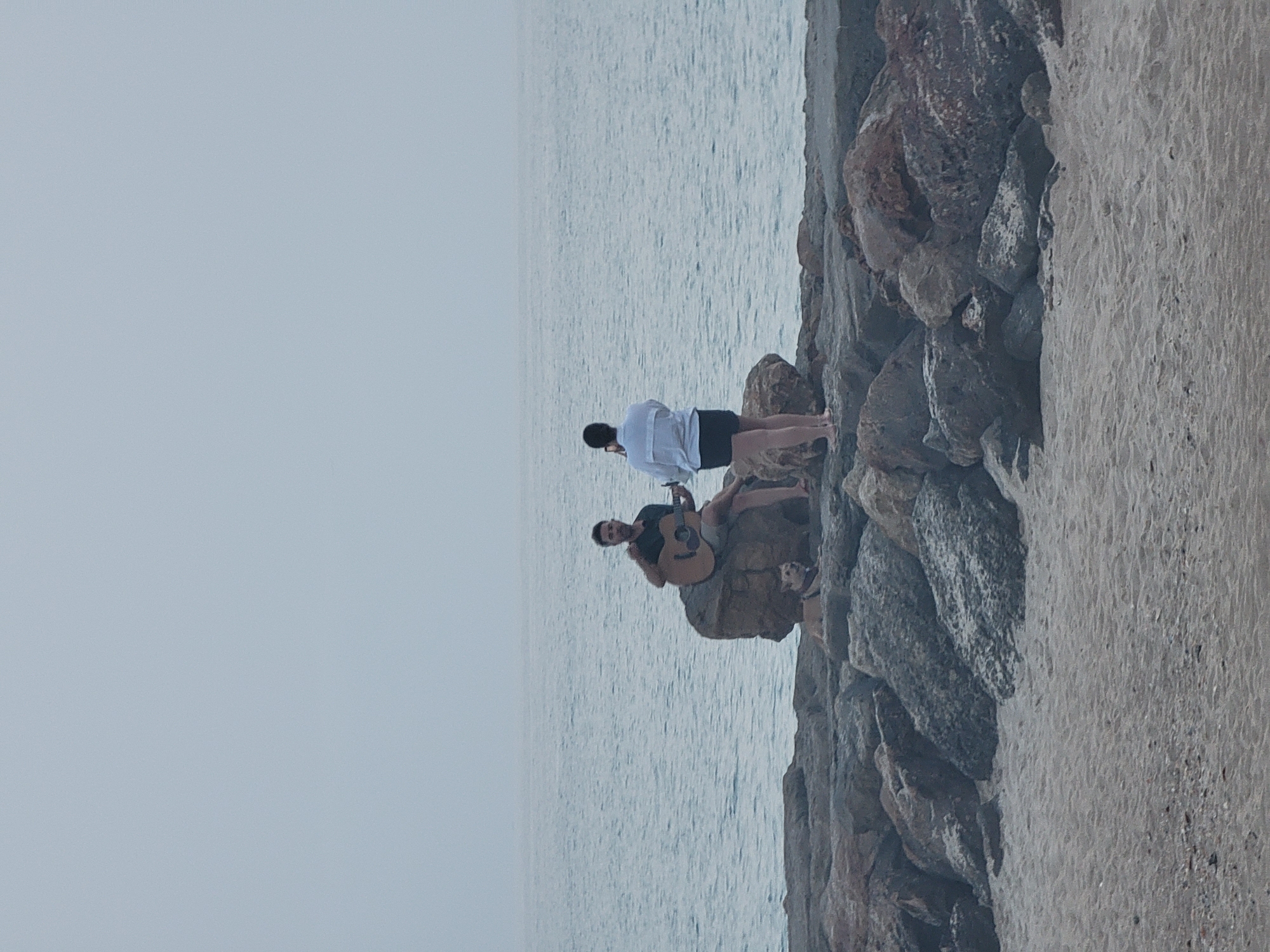 An overcast day scene depicts two people and a dog on a rocky shoreline adjacent to the Atlantic Ocean in Quarteira, Portugal. In the mid-ground, a man sits on a large rock, playing an acoustic guitar. To his right, a woman stands facing him, holding a mobile phone up, seemingly recording or taking a picture. A small dog with a light-colored coat is positioned on the rock in front of the man. The shoreline is comprised of large, irregular stones, and in the foreground, a sandy beach is visible. The water is calm, with a gentle horizon line.