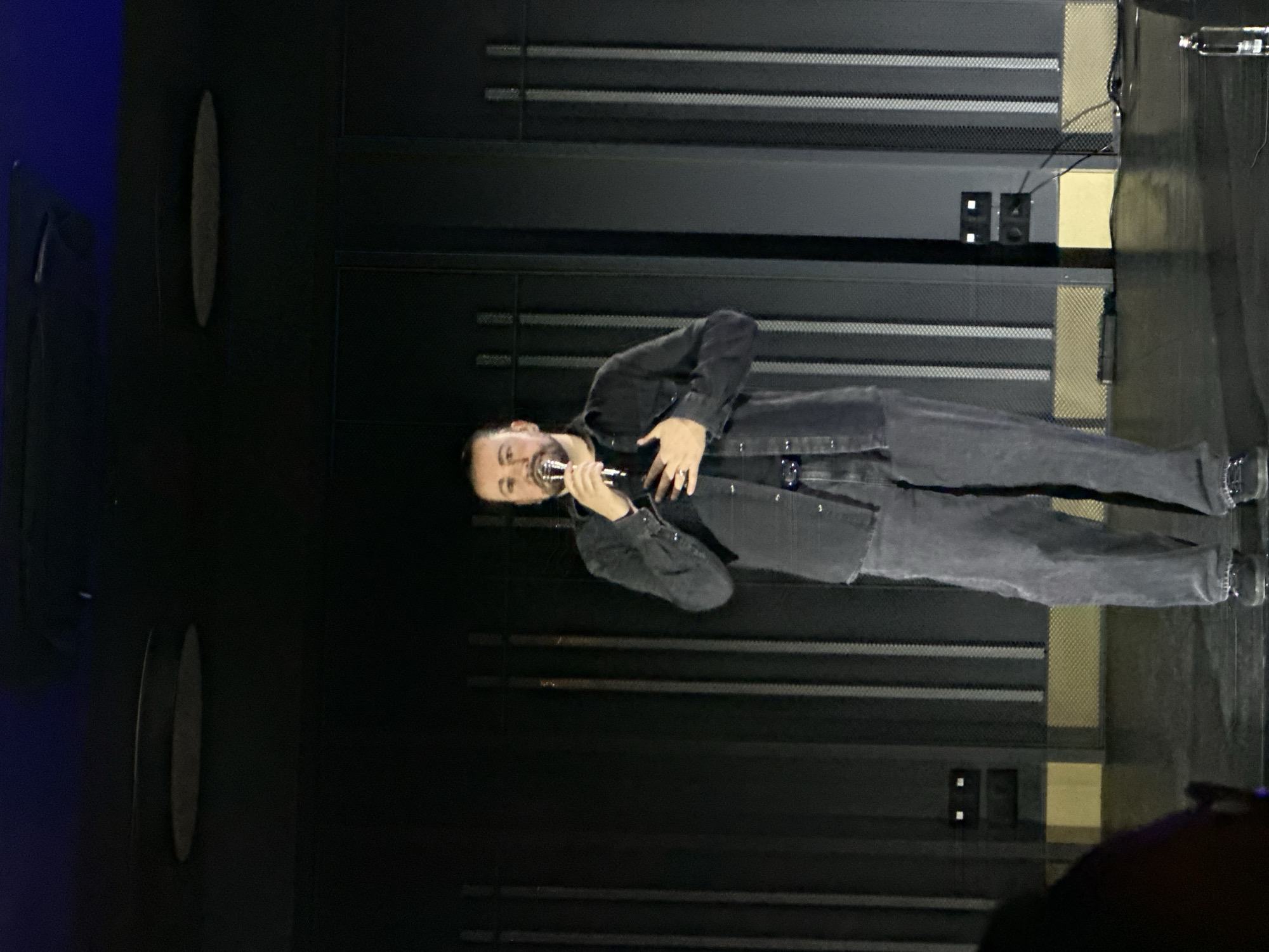 A man with a beard, wearing a dark denim jacket and pants, stands on a dark stage in Kazanlak, Bulgaria, holding a microphone and appearing to sing or speak into it. The stage backdrop consists of dark, horizontal panels with a repeating pattern. A water bottle is visible on the right edge of the frame. The lighting is dim.