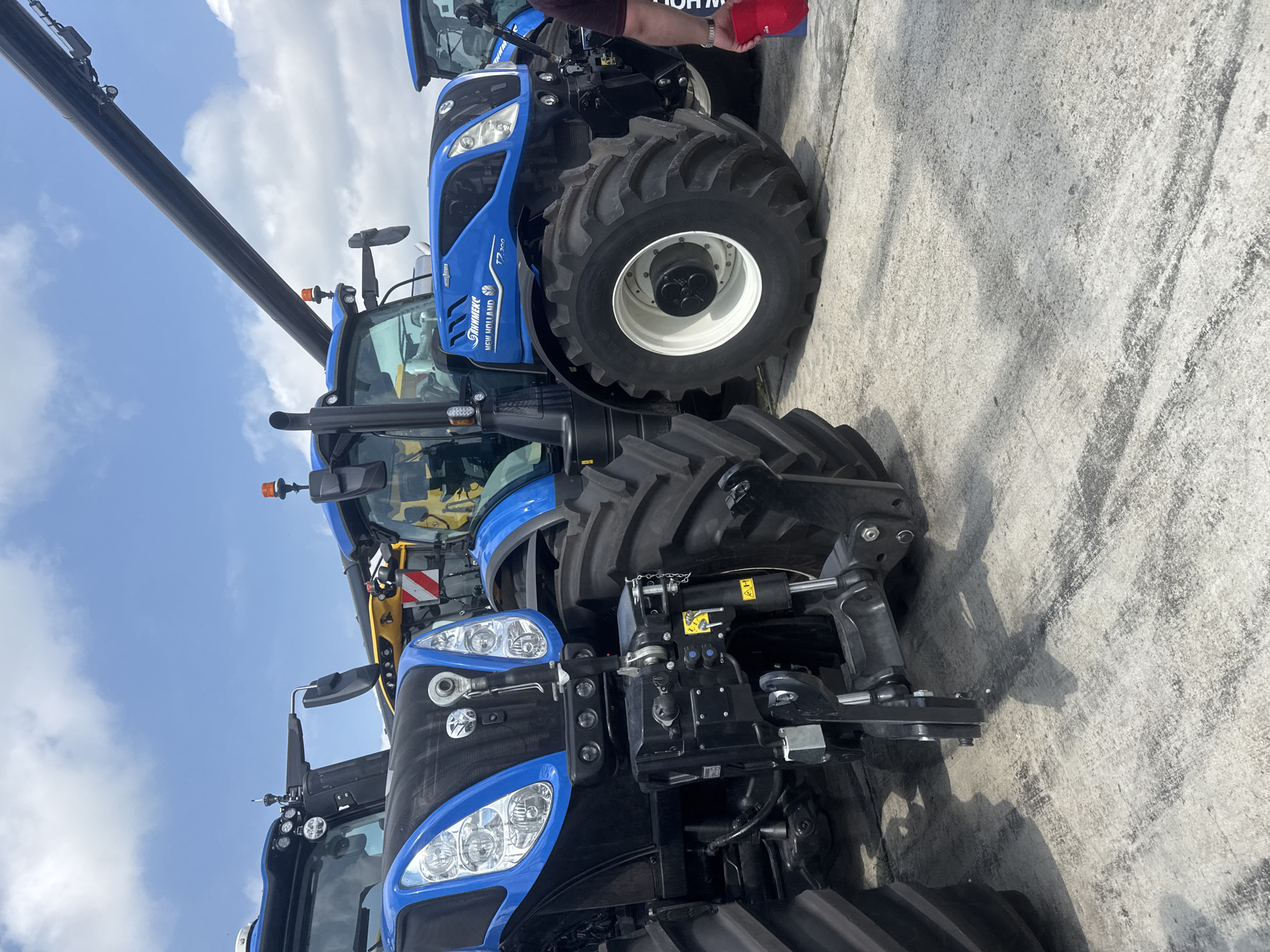 The image presents an outdoor scene featuring several large blue New Holland agricultural tractors under a bright, cloudy sky, likely at an exhibition or dealership in Stara Zagora, Bulgaria. The photograph is taken from a low angle, emphasizing the impressive scale of the machinery.
In the foreground, a blue New Holland tractor displays its front grille, multi-bulb headlights, and robust black mechanical components, possibly for a front-mounted implement. The side of the tractor clearly shows "NEW HOLLAND" and "SCHIMMELS" branding. Adjacent to it, another similar blue tractor with "T7.200" visible on its side, features a large rear wheel with "PERFORMA" marked on its tire. The ground beneath the tractors is concrete, showing some signs of wear and tire marks.
In the background, multiple other blue tractors are partially visible, suggesting a collection or display of agricultural equipment. A person's arm and a red cap are discernible near one of the background tractors, indicating human presence. A prominent, dark, articulated boom or crane arm extends diagonally across the upper left portion of the frame, silhouetted against the clear blue sky with scattered white clouds. The bright, even lighting suggests it is daytime.