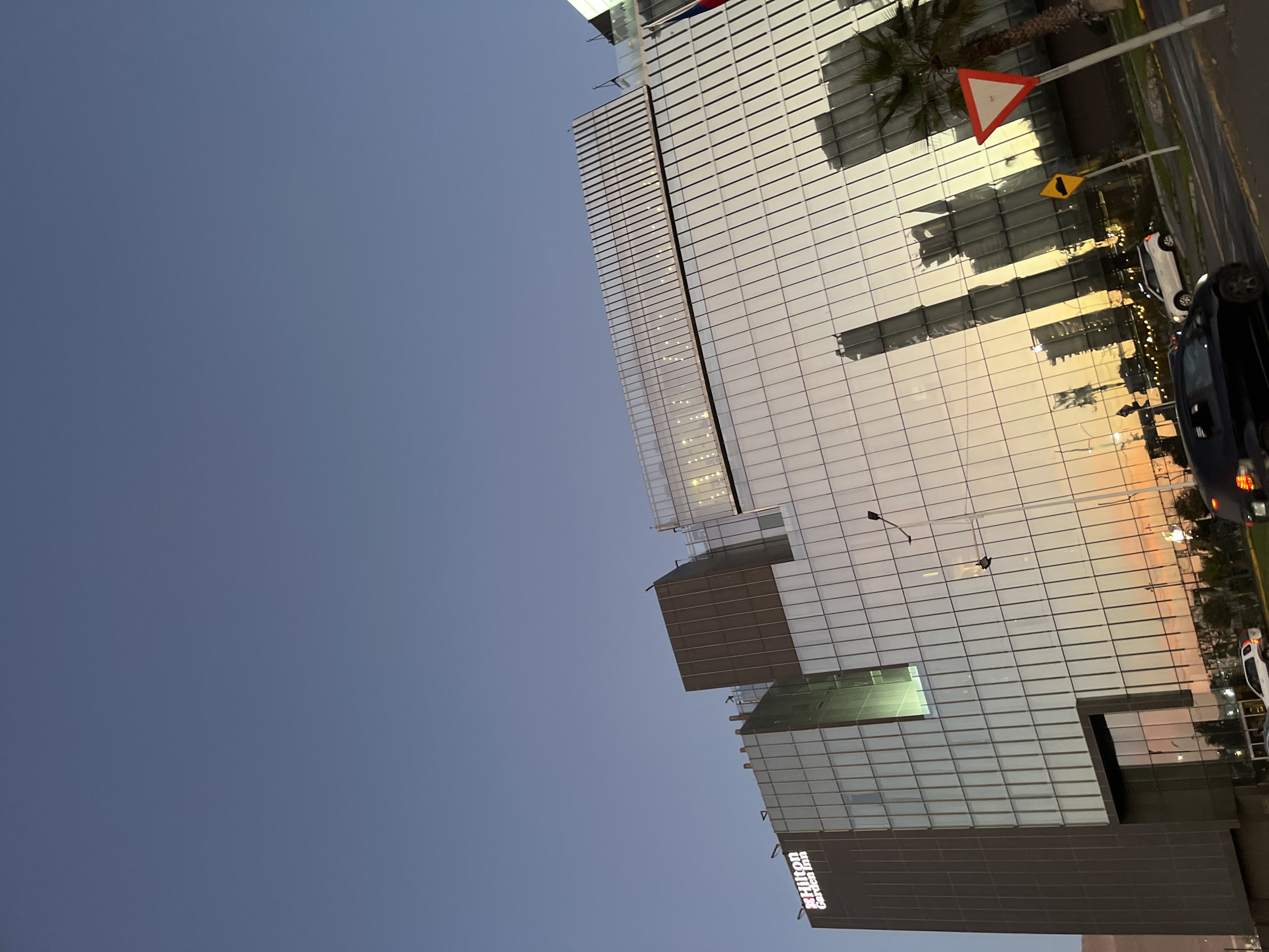 The image shows a tall, modern building with a glass facade, reflecting the surrounding city and the sky at sunset. The sky is clear and blue. The building appears to be a hotel, with "Hilton Garden Inn" visible on one side. Cars and traffic signs are visible on the street next to the building. The location is Unknown, Unknown.