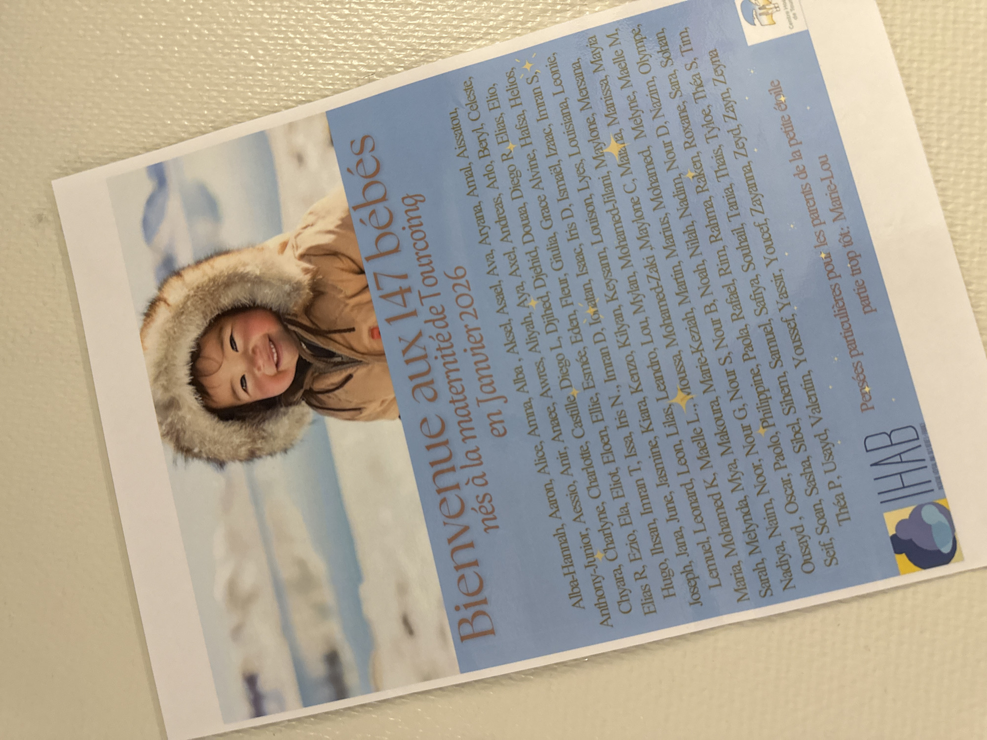 The image is a digital photograph of a flyer or poster with a blue background. At the top, there is a portrait of a smiling baby wearing a furry hooded jacket, set against a blurry background of snow and water. Below the baby's photo, text reads "Bienvenue aux 147 bébés nés à la maternité de Tourcoing en Janvier 2026". The remainder of the flyer lists numerous names, presumably of the babies born. Towards the bottom, there is text that reads "Pensées particulières pour les parents de la petite étoile partie trop tôt: Marie-Lou". A logo for "IHAB" is visible in the bottom left corner. In the bottom right, a logo for "Centre Hospitalier de Tourcoing" is partially visible. The flyer is placed on a textured surface, possibly a wall or table, with a slight sheen.