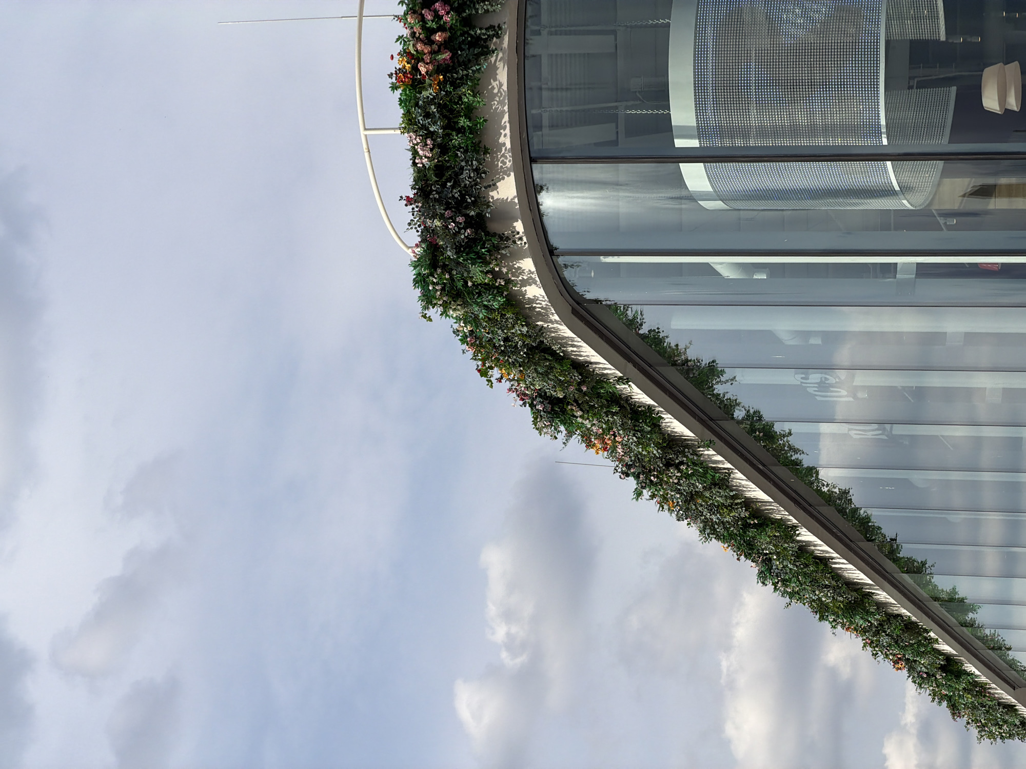 This is an image of a curved building with a large glass facade. The building is decorated with a garland of greenery and flowers that follows the curve of the building's exterior. The garland consists of dark green foliage and small pink, white, and yellow flowers. Reflections of the sky and interior building elements are visible on the glass. The sky is overcast with scattered clouds. The lighting suggests it is daytime. The overall scene is an exterior view of modern architecture with decorative elements. No people are visible in the image. No text is legible in the image.