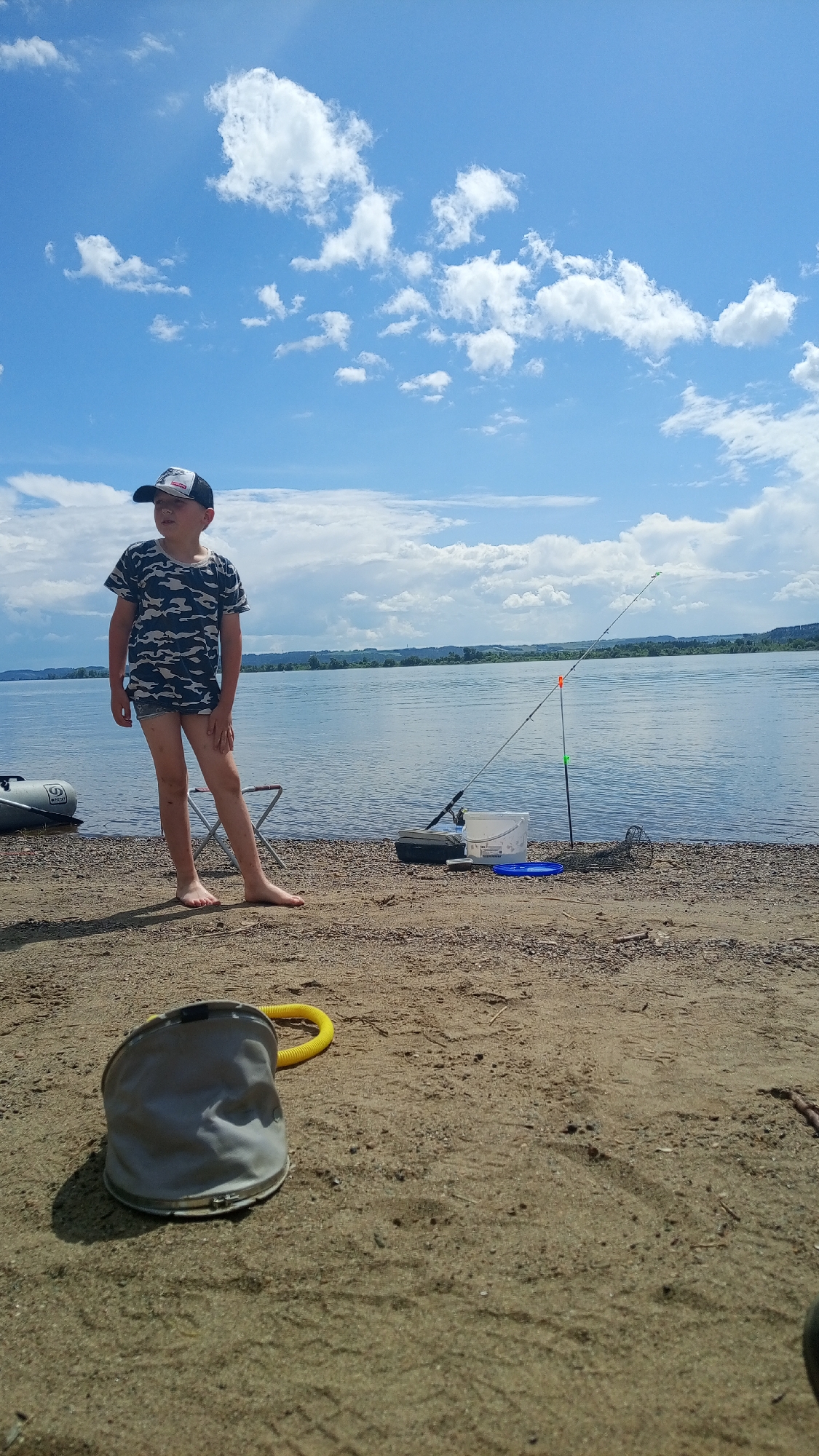 The image depicts a young boy standing barefoot on a sandy and pebbly shore beside a wide body of water in Шолья, Russia. The boy, dressed in a camouflage t-shirt, light shorts, and a backward baseball cap, is positioned slightly left of center, looking towards the water or the distant horizon under a bright, partly cloudy sky. To his right, a fishing rod is set up on a bank stick, extending over the water, accompanied by a white plastic bucket, a dark tackle box, a blue disc, and a coiled mesh fishing net. An inflatable boat is partially visible near the water's edge to the boy's left, and a small folding chair is behind him. In the immediate sandy foreground, a collapsed gray bucket with a yellow handle rests on its side. The background features a distant shoreline with faint green vegetation and low hills beneath a horizon where the sky meets the water.
