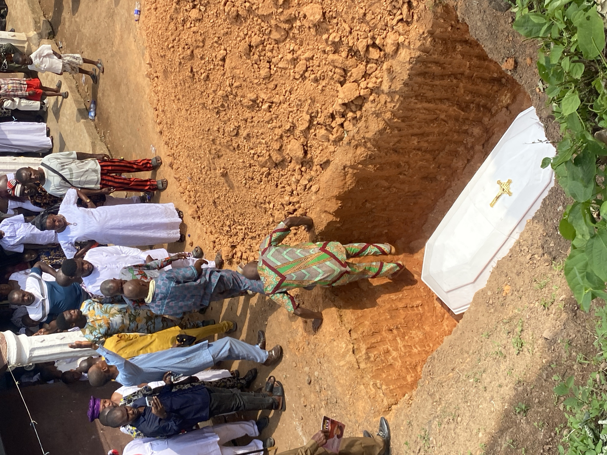 The image shows a funeral in Achara, Nigeria. A white casket with a gold cross rests in a freshly dug grave. People are gathered around the gravesite, dressed in various attire, including traditional clothing and Western-style clothes. The ground is mostly dirt and soil, with some vegetation visible at the edge of the grave.