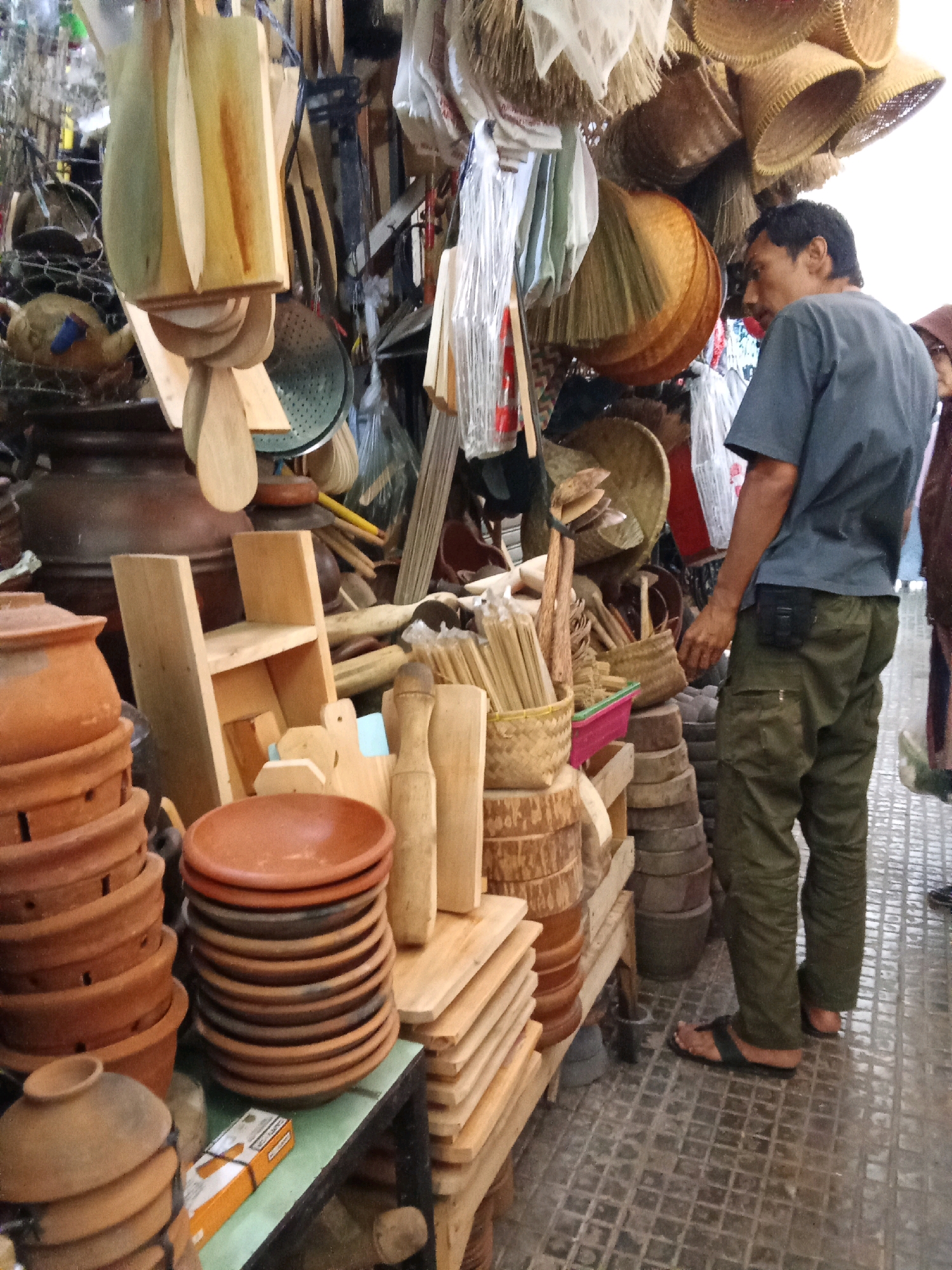 This is a medium-shot, eye-level, outdoor photograph of a stall selling various handicraft items in Banjarnegara, Indonesia. The stall displays an assortment of wooden utensils, ceramic pots and bowls, woven baskets, and bamboo items. In the foreground, stacked terracotta pots and a pile of stacked terracotta plates are visible. To the right, a man wearing a grey t-shirt and olive green cargo pants stands facing left, examining the merchandise. He is wearing sandals. The ground is paved with square tiles. The background reveals more of the stall's inventory, including more woven baskets hanging from above. The overall impression is a bustling market scene.