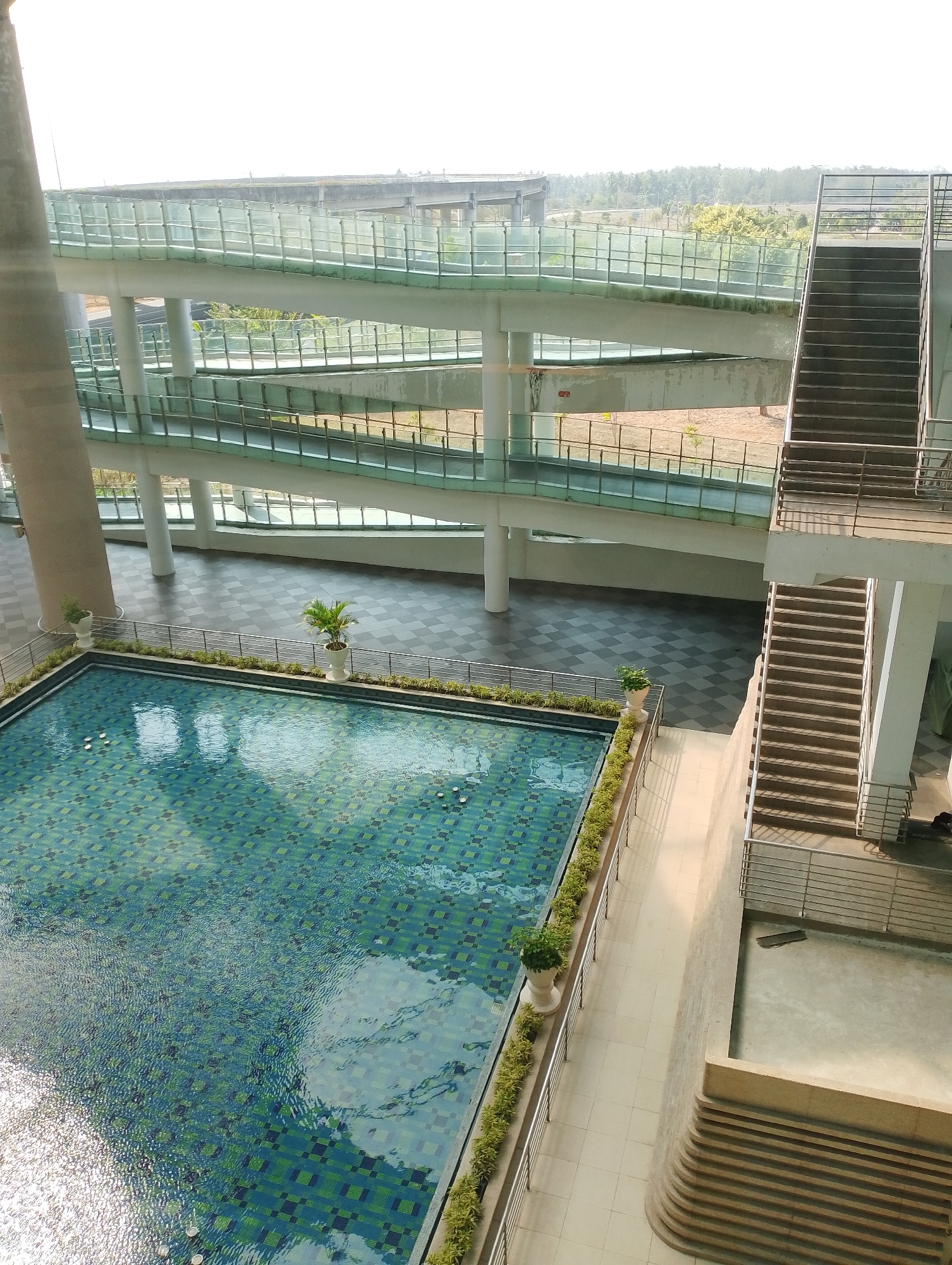 This is an aerial, eye-level shot of a modern, multi-level walkway and a swimming pool, likely located in Glagah, Indonesia, judging by the surrounding landscape and architecture. The walkway features multiple ramps with clear glass barriers and metal railings, winding their way up. To the right, a set of dark grey, outdoor stairs with metal railings ascends. Below the walkways and stairs is a large, rectangular swimming pool with a blue and green tiled bottom. The pool has a rippling surface reflecting the sky and surrounding structures. Along the edge of the pool is a walkway with light-colored tiles, bordered by small green plants in white planters and metal railings. The scene is bathed in natural light, suggesting daytime. There are no visible people in the image. The overall impression is one of contemporary urban design integrated with recreational facilities.