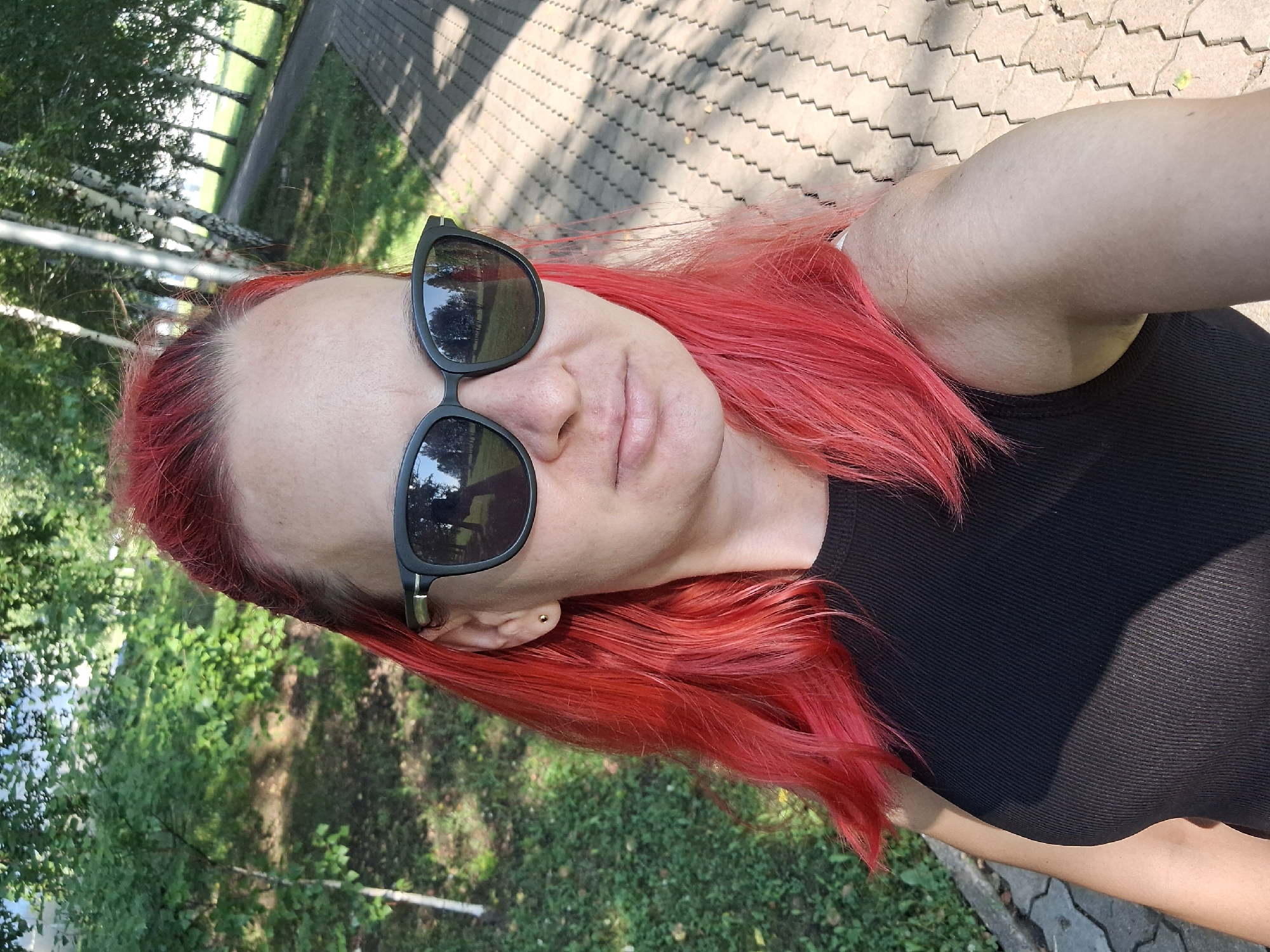 A woman with bright red hair and sunglasses is taking a selfie in an outdoor setting. She is wearing a black sleeveless top. The background shows a park-like environment with trees and a paved pathway. The lighting suggests it is daytime. The location is identified as Atamanovka, Russia.