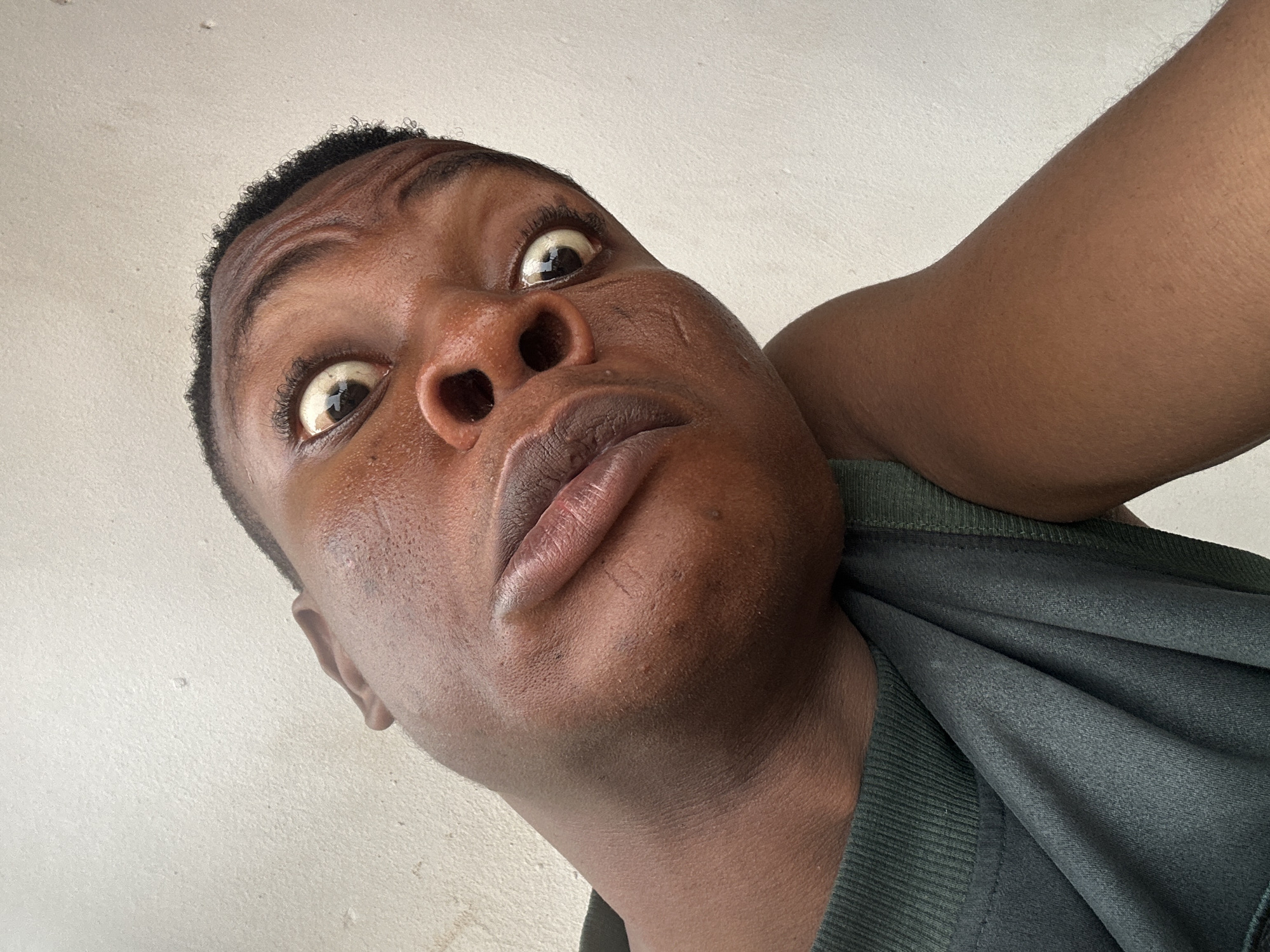 A low-angle close-up of a Black man with wide eyes, looking upwards at the camera. He is wearing a dark green t-shirt. The background is a light, off-white wall. The location in the city is Unknown, Unknown.
