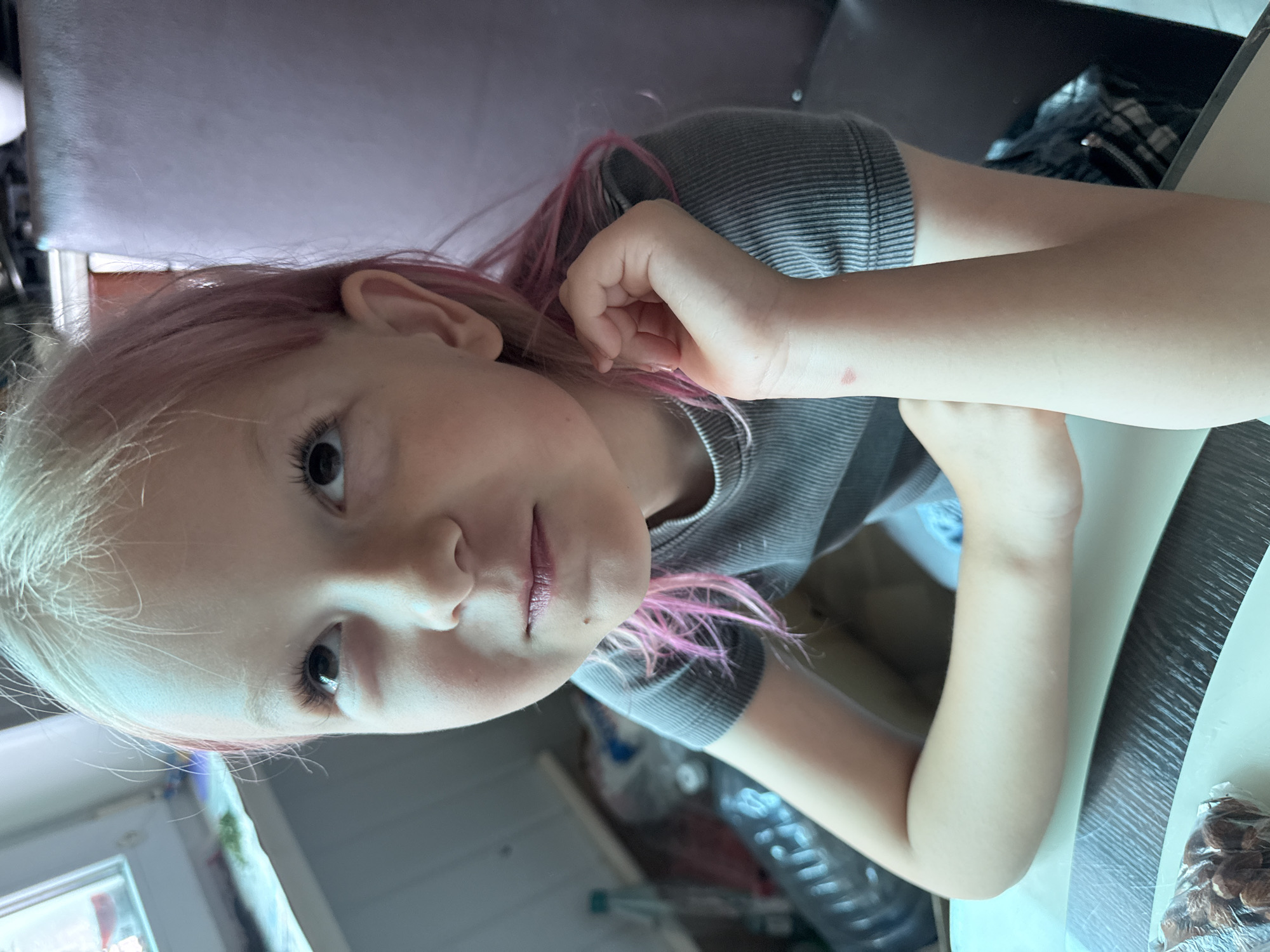 An overhead medium shot captures a young girl with blonde hair streaked with pink, looking upwards with her right arm resting on her chin. She wears a grey, ribbed t-shirt. The background is blurred, showing a window on the left with items on a windowsill, and a dark chair on the right. The girl is seated at a table with a white placemat and a plastic bag of nuts. The lighting suggests an indoor setting, likely during the day.