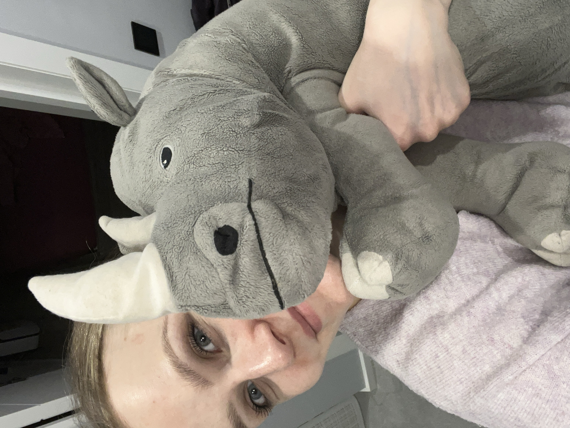 This media file, taken in Khimki, Russia, features a young woman interacting with a large, gray, plush rhinoceros toy indoors. The image is oriented sideways, suggesting the subjects are lying down or the camera was held horizontally.
The woman's face and one hand are visible. She has light-colored hair, fair skin, and blue-gray eyes with visible eyelashes and eyebrows. Her expression is neutral. Her left arm is wrapped around the body of the plush rhinoceros, indicating she is holding or cuddling it. She is partly covered by a light purple, knitted garment, likely a sweater or blanket.
The plush rhinoceros is a prominent subject, characterized by its soft, textured gray material, white horn, visible ears, black button-like eyes, and a stitched black mouth and nostril. The background shows portions of a white surface and a dark area, consistent with an indoor setting like a bedroom. There is no visible text in the image.