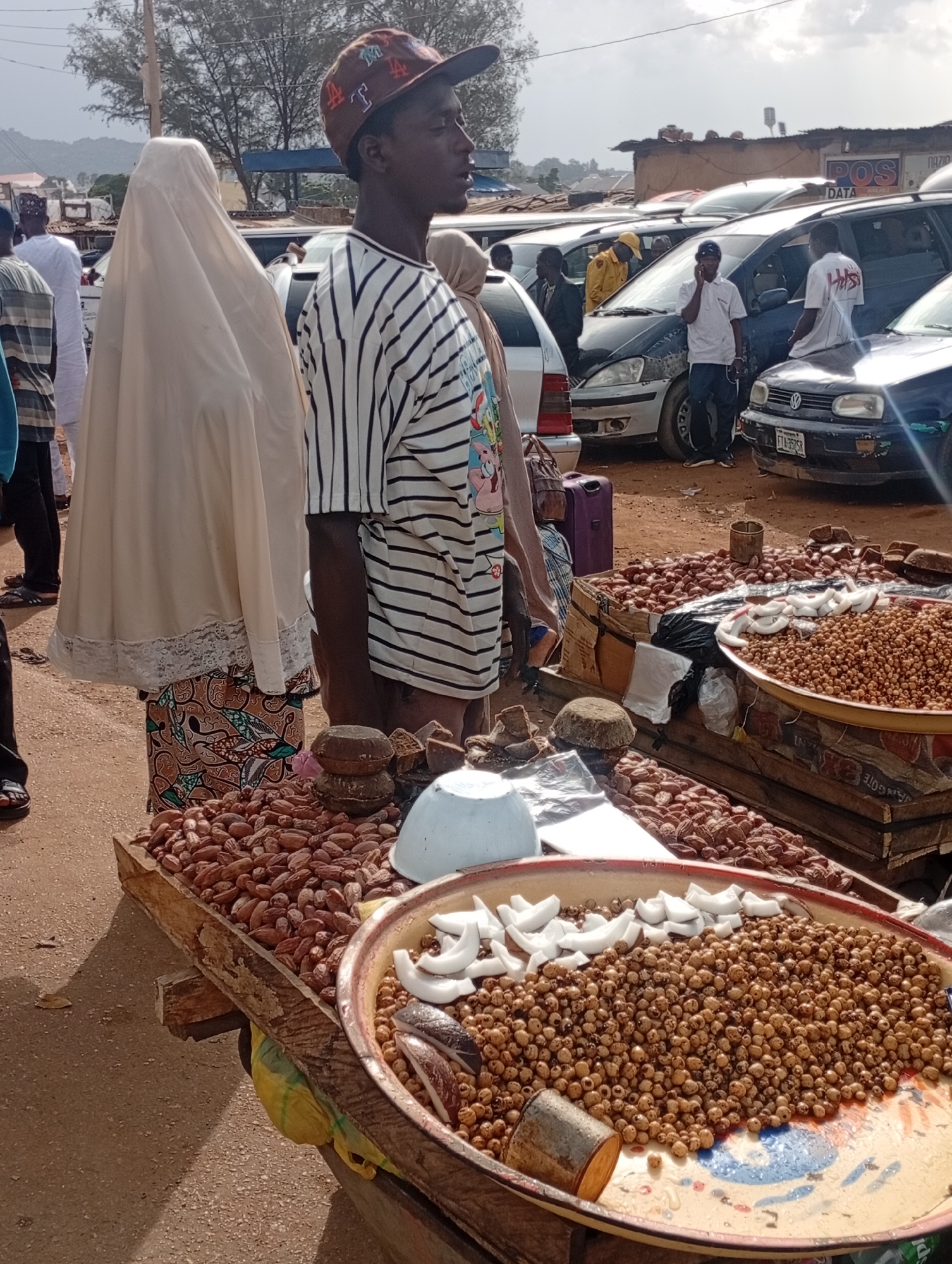 In a bustling outdoor market in Rafin Par, Nigeria, a vendor stands behind a display of nuts and dried fruits. The vendor, a young man wearing a striped t-shirt and a baseball cap, looks to his right, his gaze fixed on something out of frame. Beside him, a woman in a flowing white hijab and a patterned skirt, her face hidden, also appears to be waiting.
The market scene is filled with activity. Cars are parked in the background, suggesting a busy thoroughfare or a busy shopping area. Various goods are laid out on makeshift stalls. In the foreground, the vendor's wares are prominently displayed in wooden crates and a large, colorful bowl. The bowl overflows with small, round nuts, likely tiger nuts, topped with slices of white coconut. Another crate is filled with larger, oval-shaped dried fruits, possibly dates or prunes.
The time of day appears to be late morning or early afternoon, with bright sunlight casting shadows. The sky is partly cloudy, hinting at a warm, possibly humid, day. The ground is dusty, typical of a market in this region. Signs are visible in the background, including one that reads "POS DATA," indicating a point-of-sale transaction service is available. The overall atmosphere suggests a typical day at a Nigerian market, characterized by commerce, people going about their daily business, and an array of local produce.