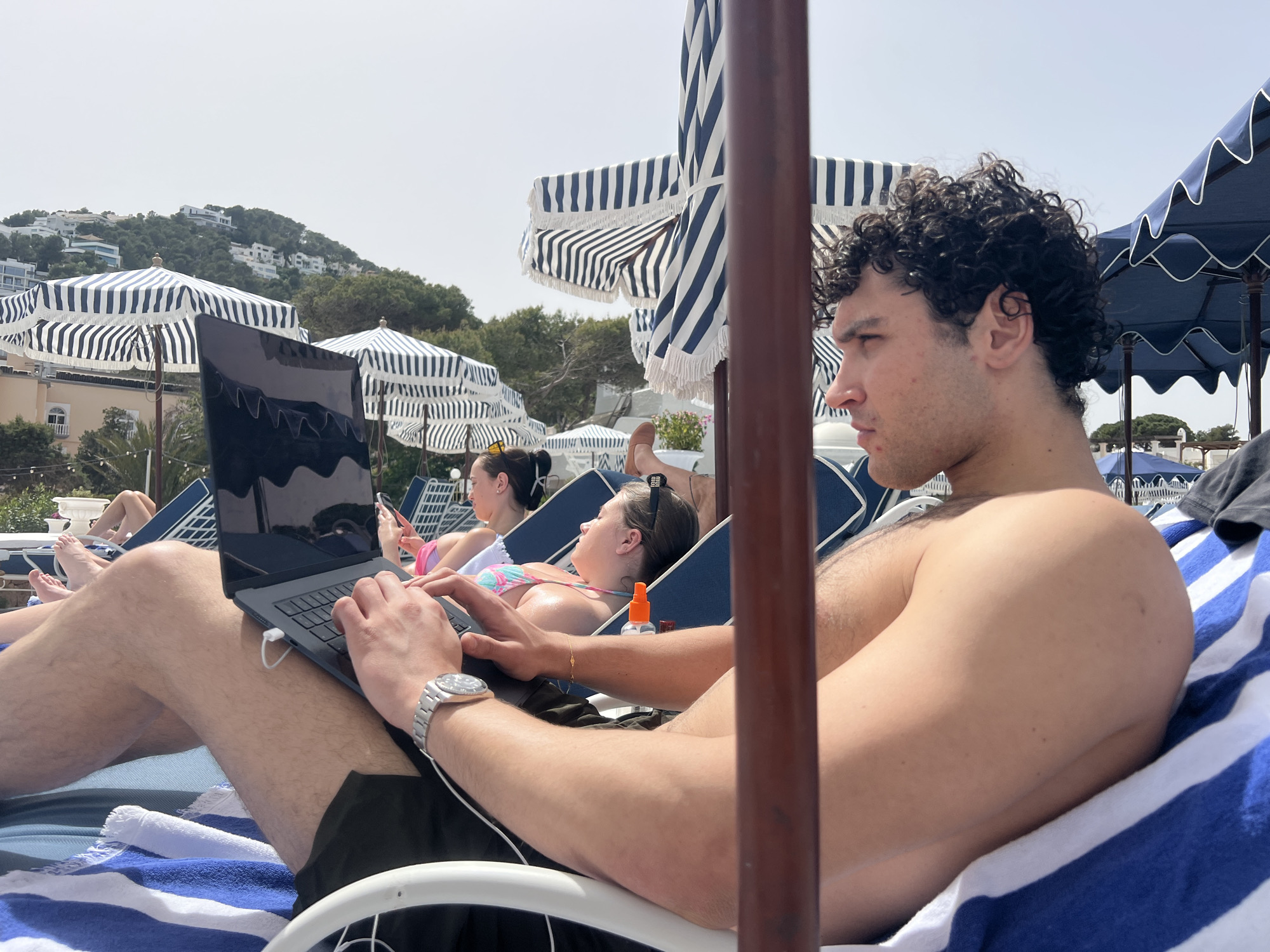 The image shows a man using a laptop while lying on a blue and white striped lounge chair under a blue and white striped beach umbrella. He is shirtless and wearing a wristwatch. In the background, other people are sunbathing on similar lounge chairs, with additional beach umbrellas visible. Buildings are seen on a hillside in the distance. The location is likely Urbanització Siesta, Spain, based on visual cues, although the image itself lacks specific landmarks to confirm this.