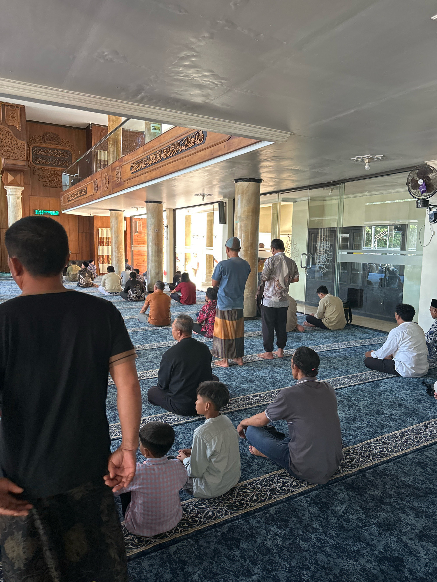 The image depicts the interior of a mosque in Malang, Indonesia, where a group of men and boys are seated on a blue carpeted floor, facing the front of the mosque. The mosque features traditional Islamic architecture with wooden paneling and decorative carvings on the walls and a mezzanine level. Natural light streams in from large windows, illuminating the space. Several men are seen praying or observing the prayer in unison, indicating a religious gathering. The overall atmosphere is one of reverence and community.