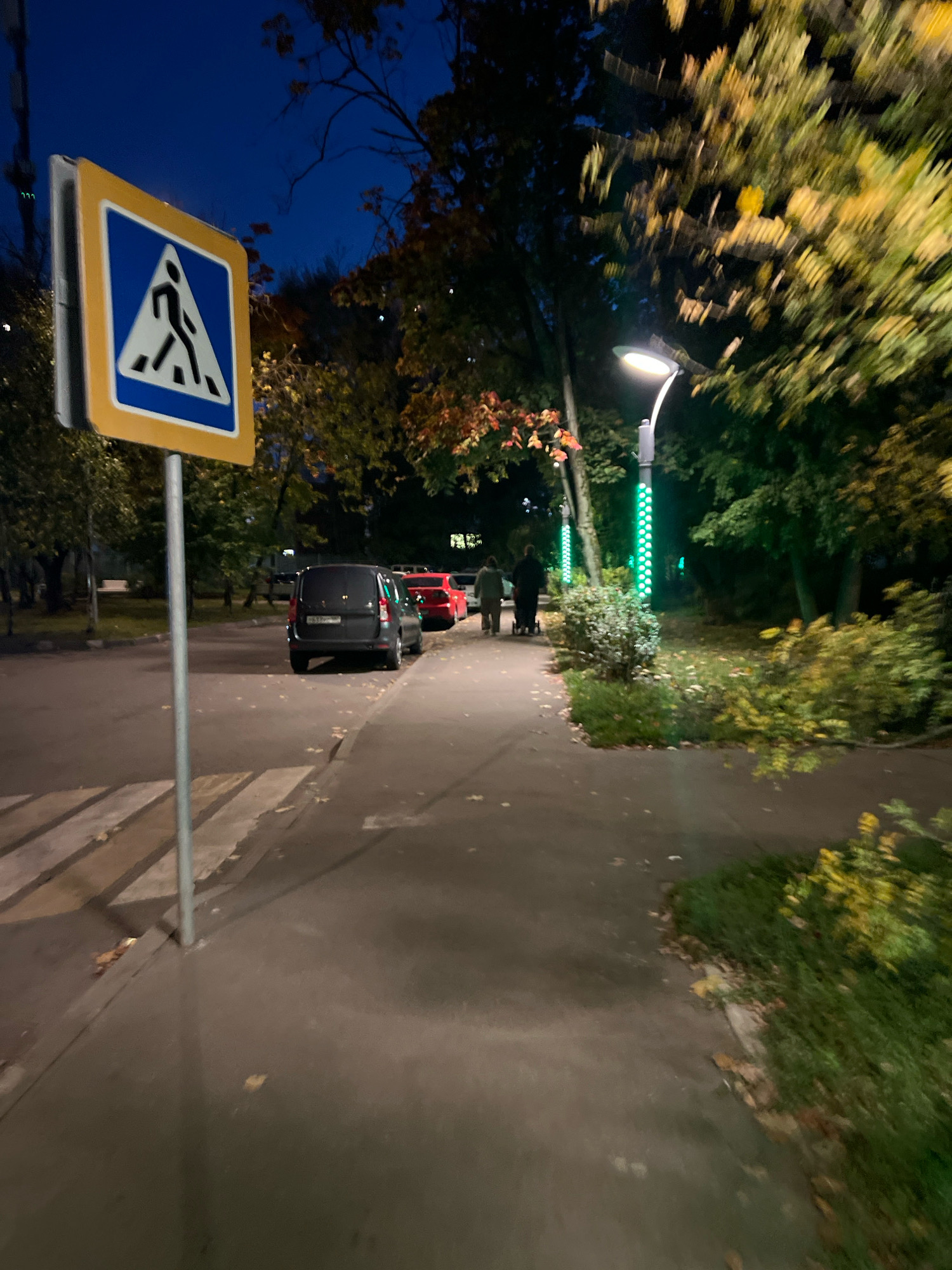 The image depicts a nighttime scene on a sidewalk in Moscow, Russia. A pedestrian crossing sign is prominently displayed on the left. In the background, several cars are parked along the street, with a red car and a dark-colored van being most visible. Two individuals are walking away from the camera, one pushing a stroller, on the sidewalk near a lamppost with green lights. Trees with autumn foliage line the area, suggesting the season is fall. The sky is dark blue, indicating it is evening or night. The ground is paved with asphalt and concrete, and fallen leaves are scattered on the walkway.