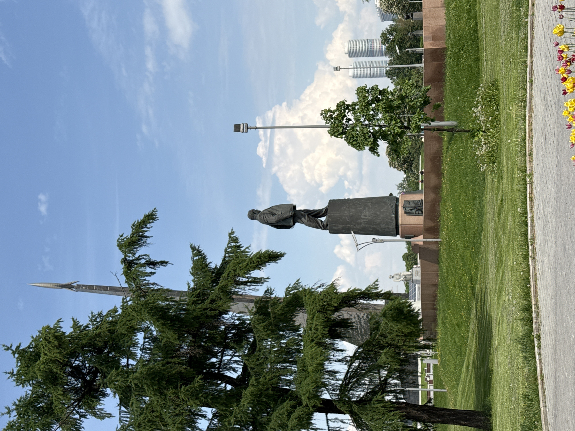 The image shows a statue of a man on a pedestal in a park-like setting. The statue is of a man in a suit-like outfit, standing atop a dark-colored pedestal. Behind the statue is a blue sky with fluffy white clouds. To the left of the statue is a large coniferous tree, partially obscuring a tall, spire-like structure. In the background, there are buildings, including some tall, cylindrical structures. The park area includes green grass and flowers, with a concrete walkway on the right side. The location is likely in Moscow, Russia, given the architectural style of the buildings and the presence of a monument reminiscent of Soviet-era designs.
