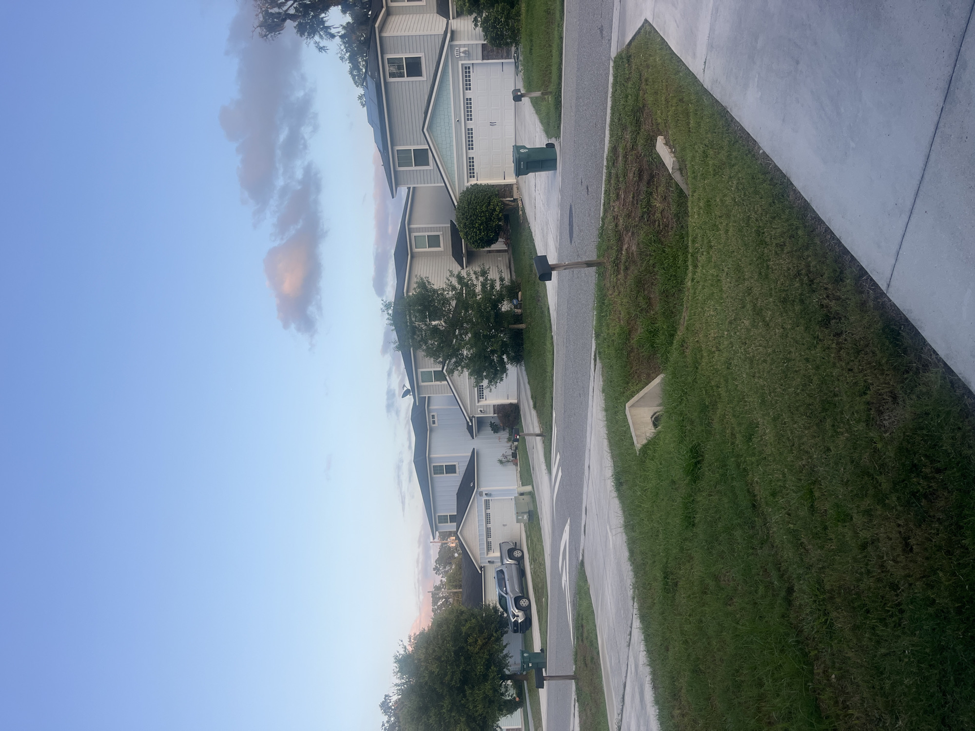 The image depicts a suburban street scene in Jacksonville, Florida, United States of America, during early morning or late evening with a clear sky and soft pastel-colored clouds. The foreground features a concrete sidewalk and a grassy area with a drainage culvert. In the middle ground, a paved road curves through a residential neighborhood lined with modern two-story houses. These houses are painted in light blue and gray tones, with white trim and garages. A silver pickup truck is parked in a driveway. Green trash bins are visible on the sides of the road. The background shows more houses and trees, with the sky taking up the upper portion of the frame. The overall impression is one of a quiet, well-maintained residential area.